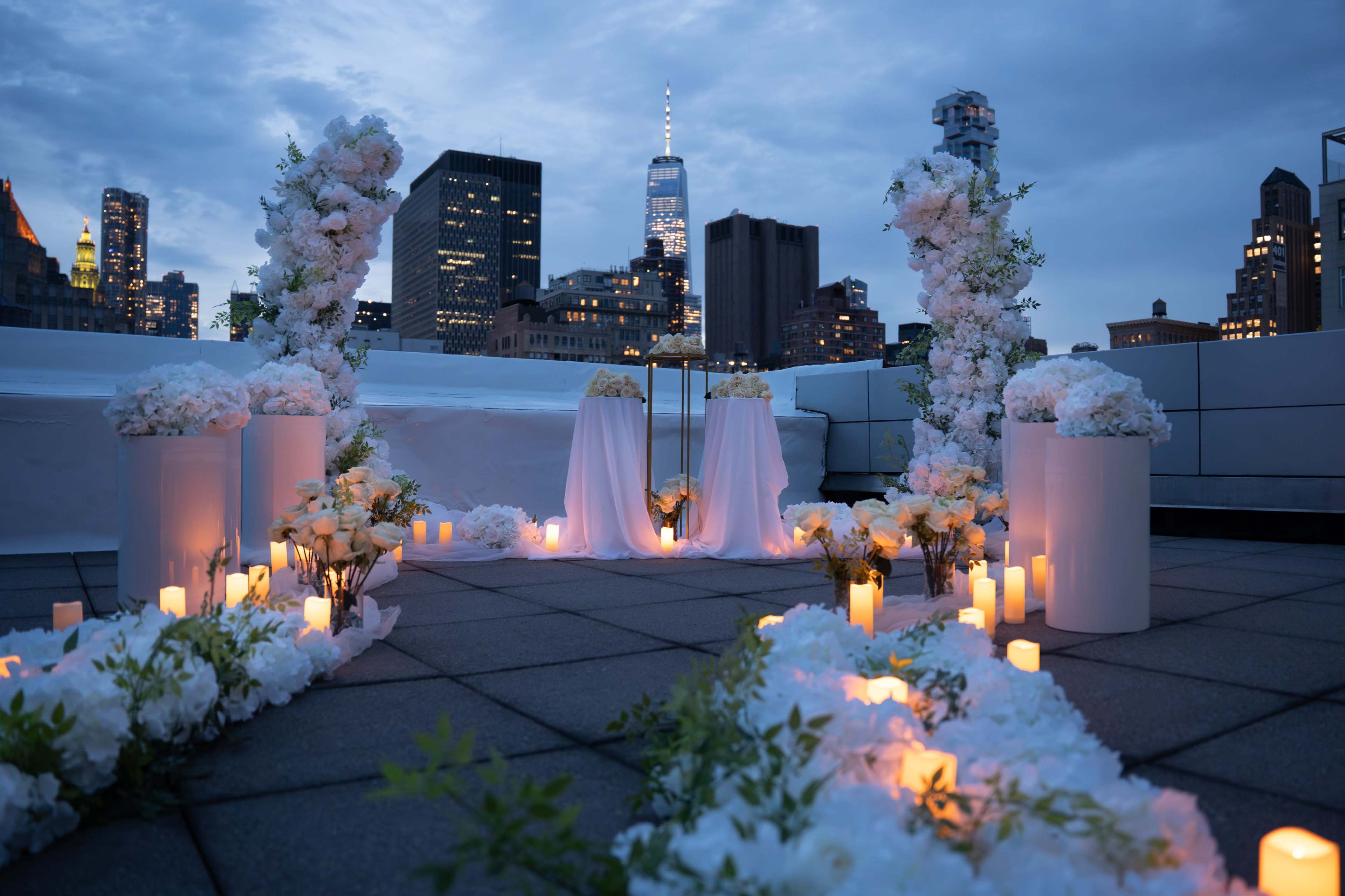 A rooftop ceremony setup is displayed with floral arrangements, candles, and a backdrop of a city skyline at dusk.