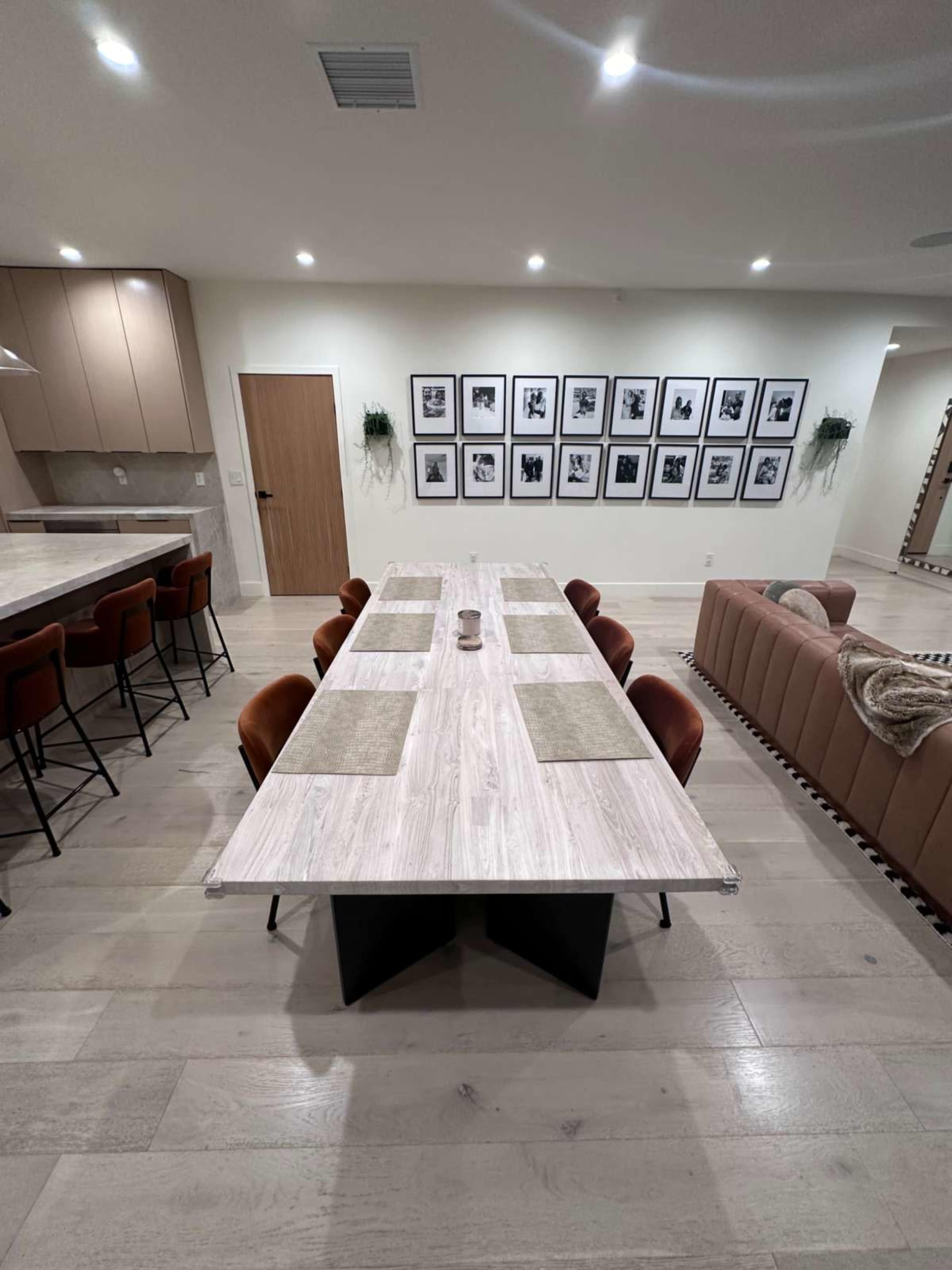 The image shows a modern dining area with a long, rectangular table surrounded by eight chairs, a gallery wall of framed photos, and a sofa in the adjacent space.