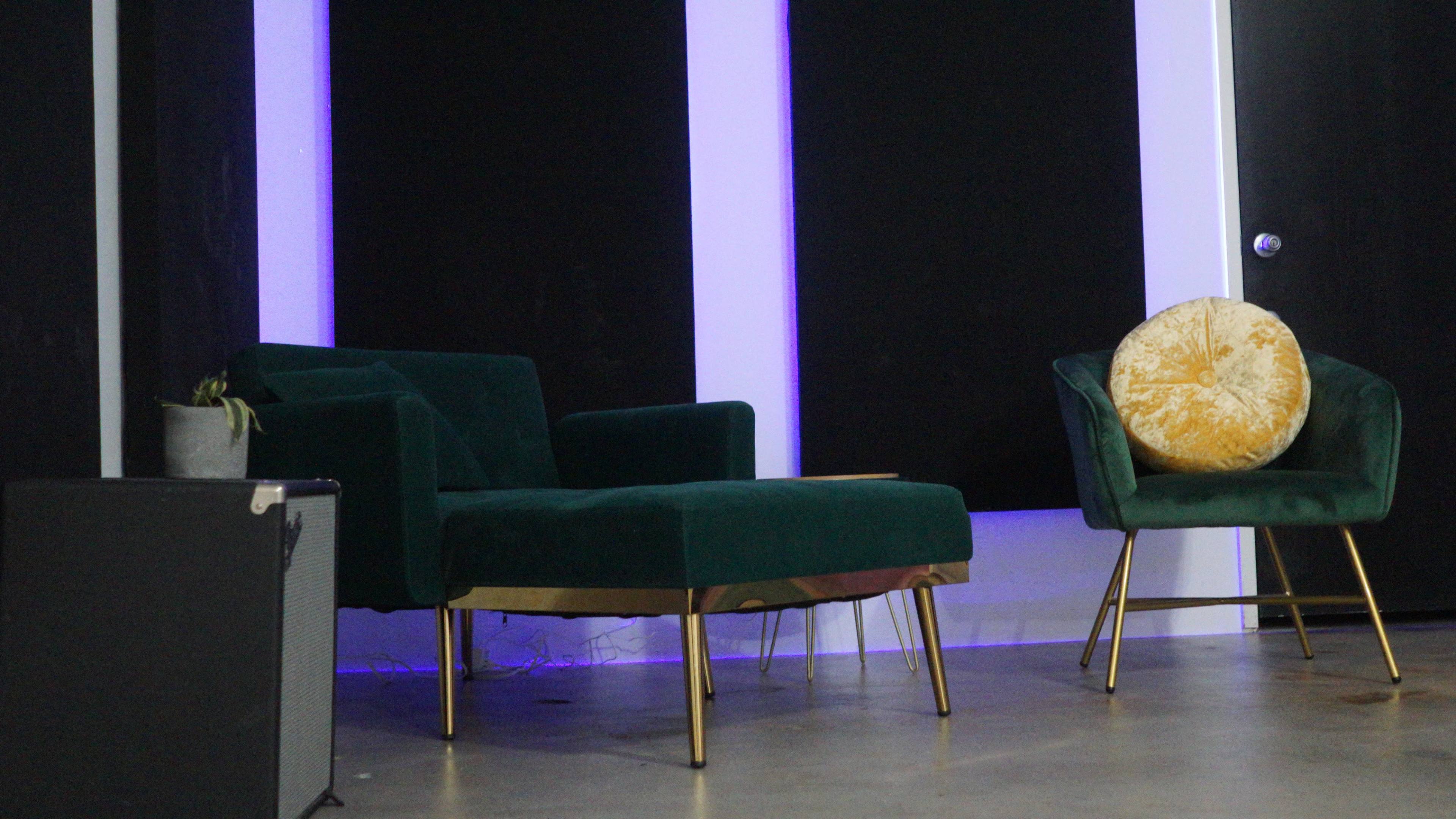 A minimalistic interior featuring two green armchairs, a round decorative pillow, and a black speaker in front of a backdrop with purple lighting.