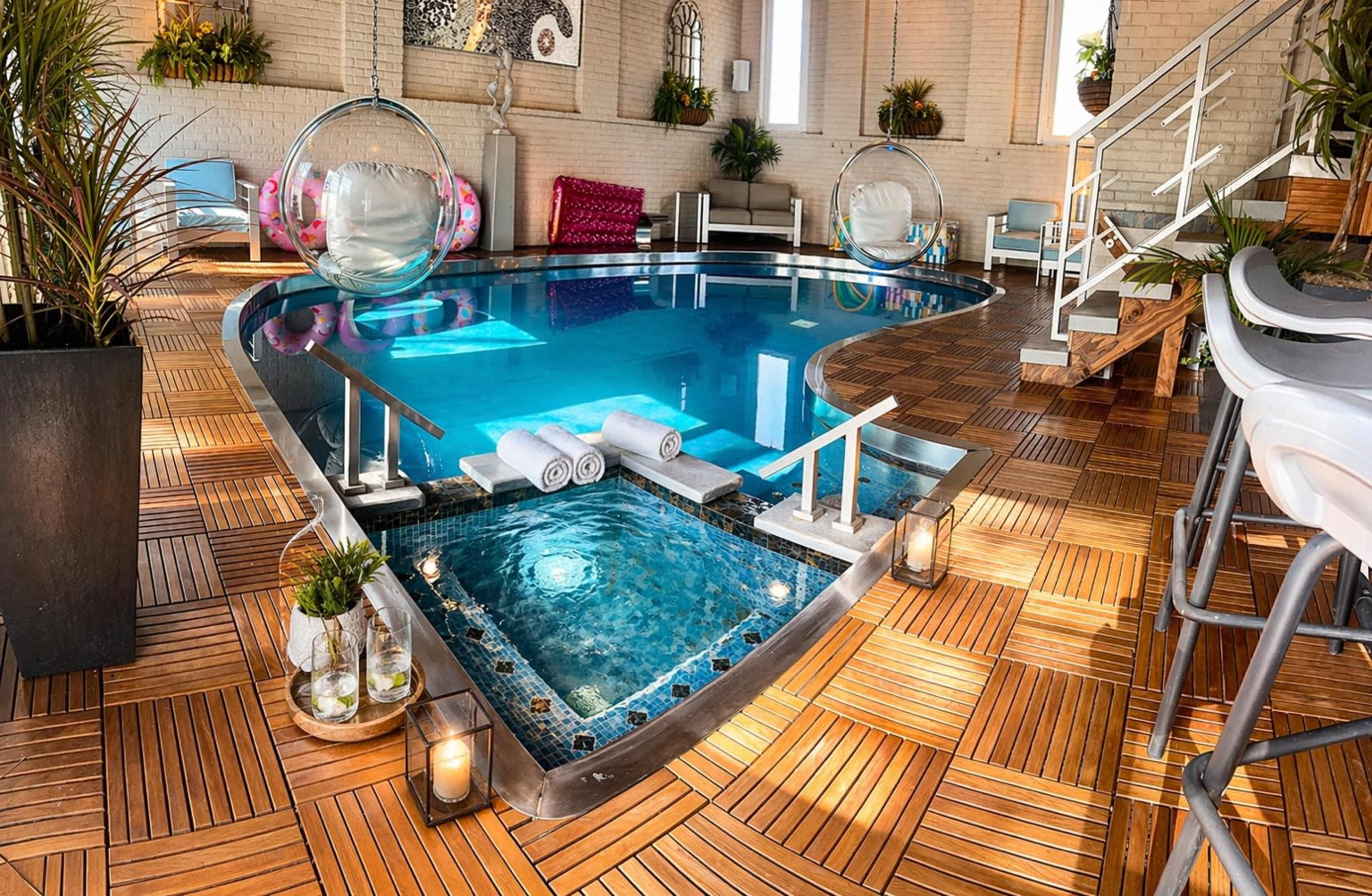 The image shows a modern indoor pool area featuring a large swimming pool, a smaller whirlpool, and elegant wooden decking, surrounded by decorative plants and seating.