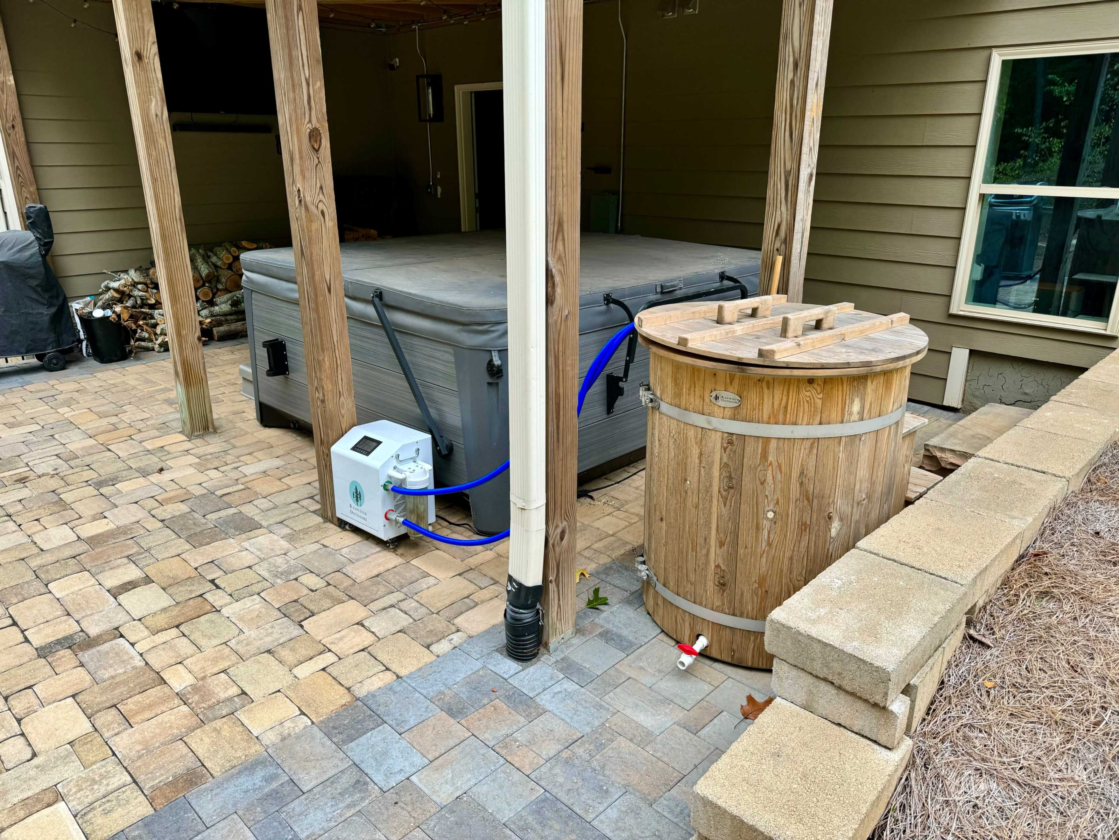 A hot tub and a wooden barrel on a paved patio next to a house.