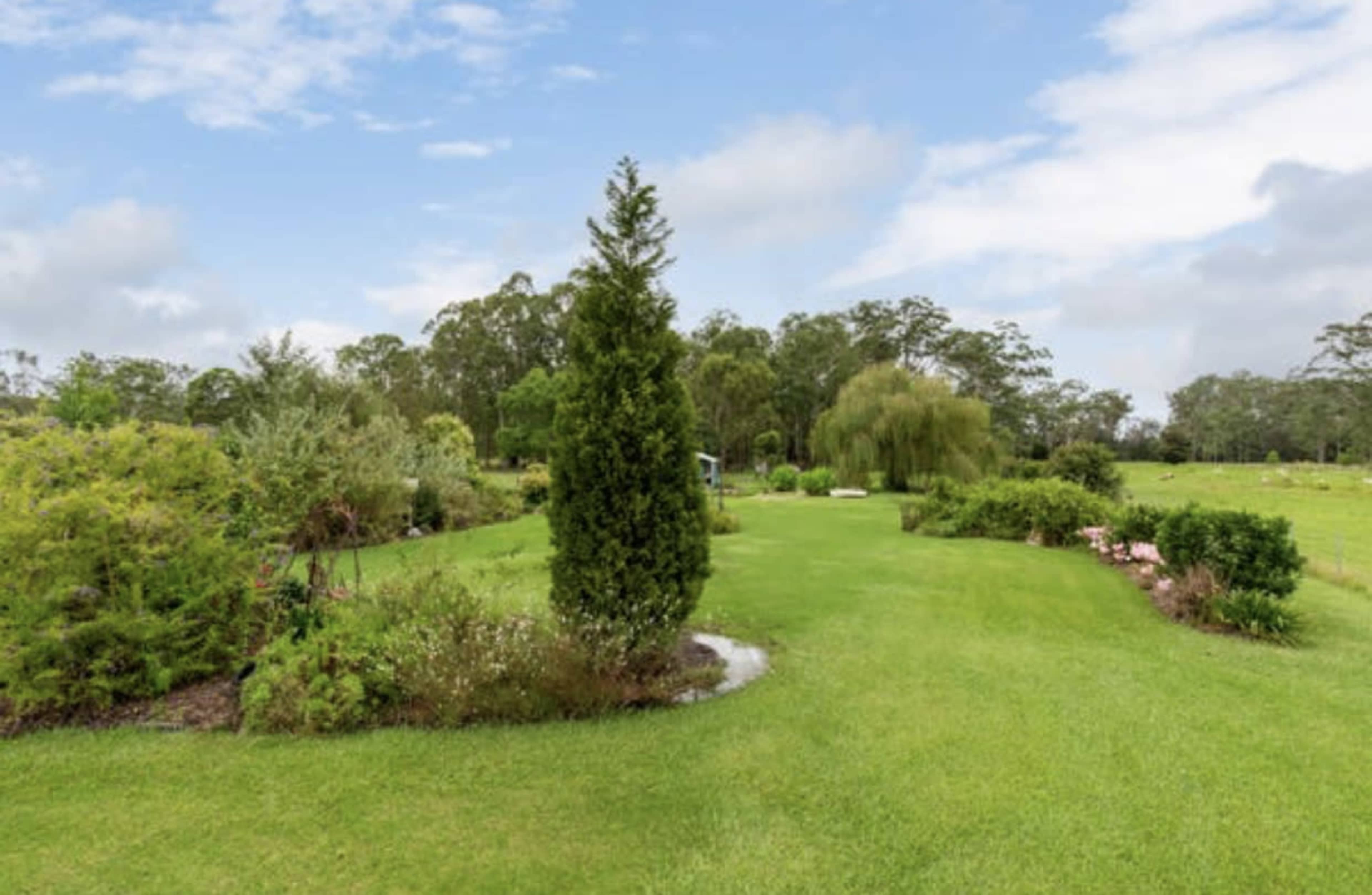 The image shows a lush garden with neatly trimmed grass, various plants, and trees under a partly cloudy sky.