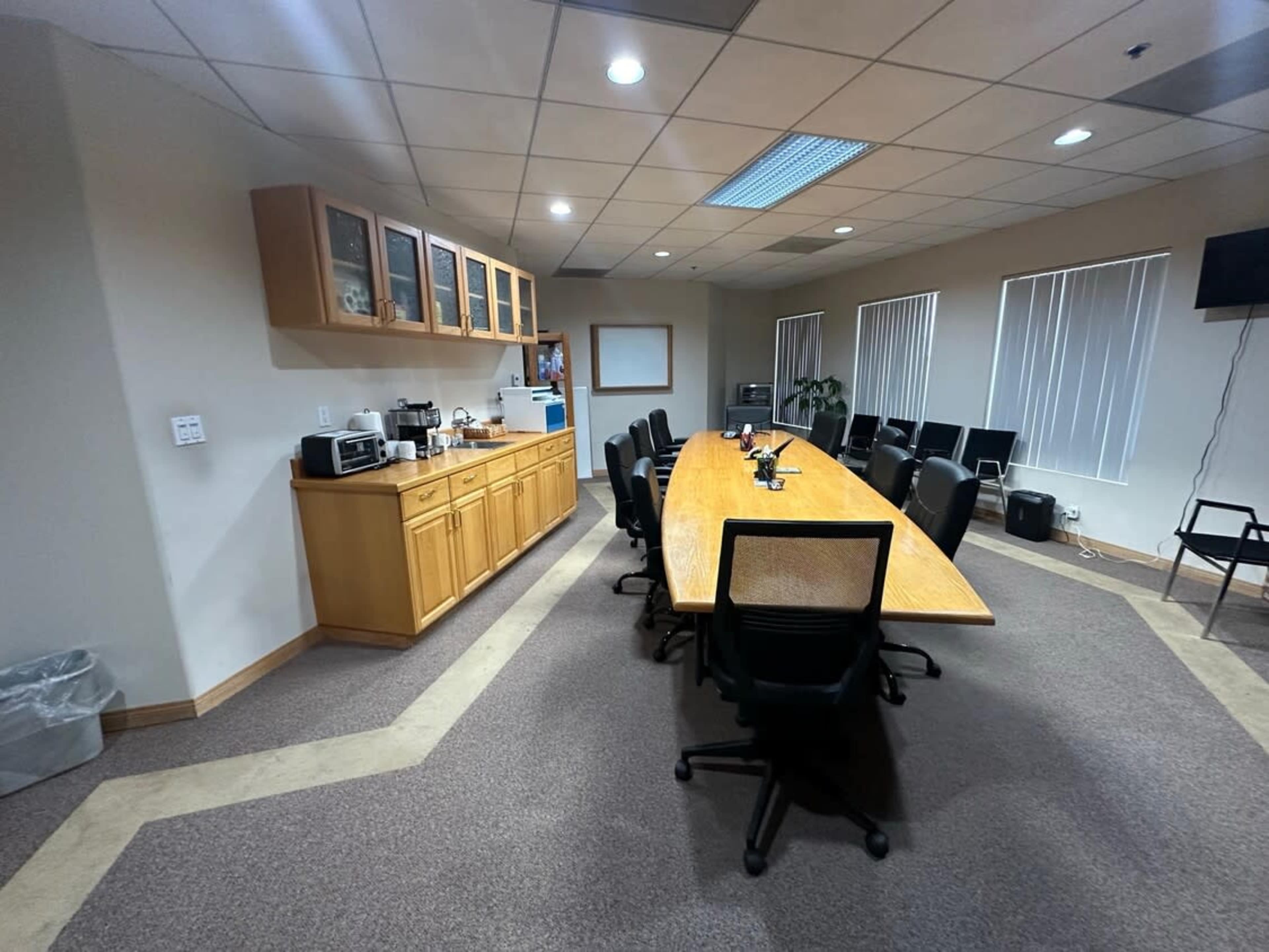 The image shows a conference room equipped with a long wooden table, black chairs, and a kitchenette area along one side.