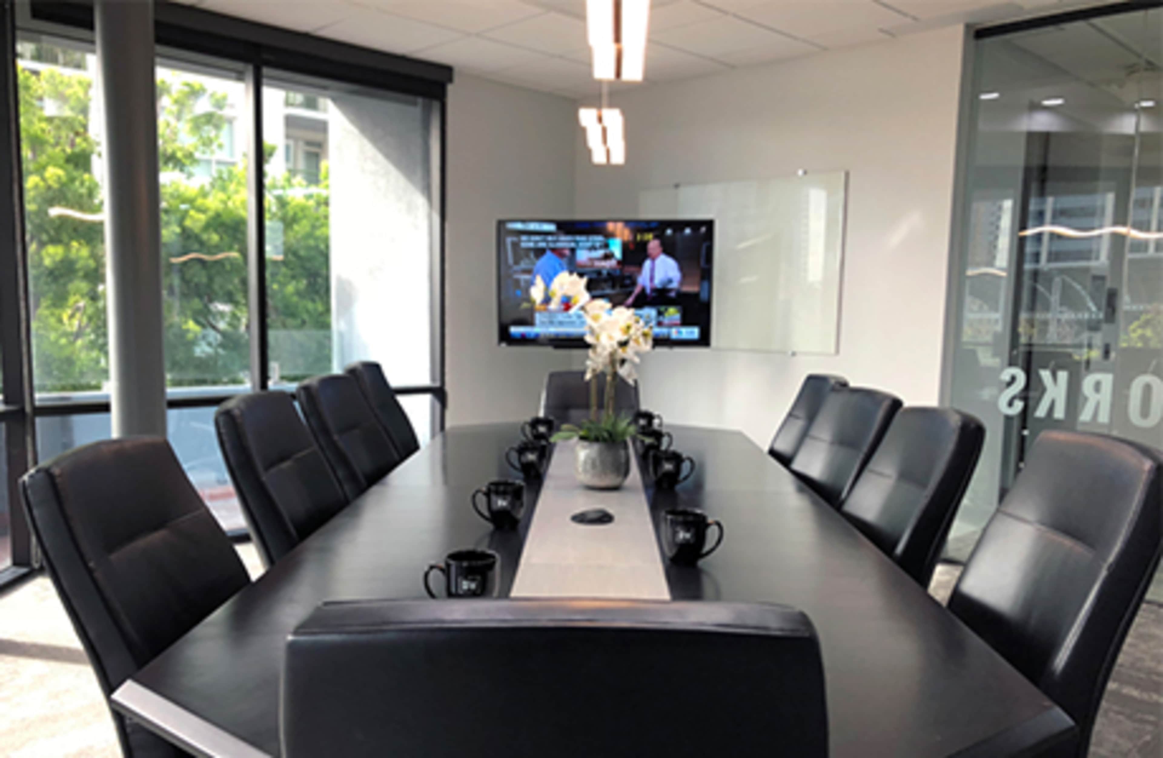 A modern conference room features a large table surrounded by black chairs, with a television screen displaying information on the wall.
