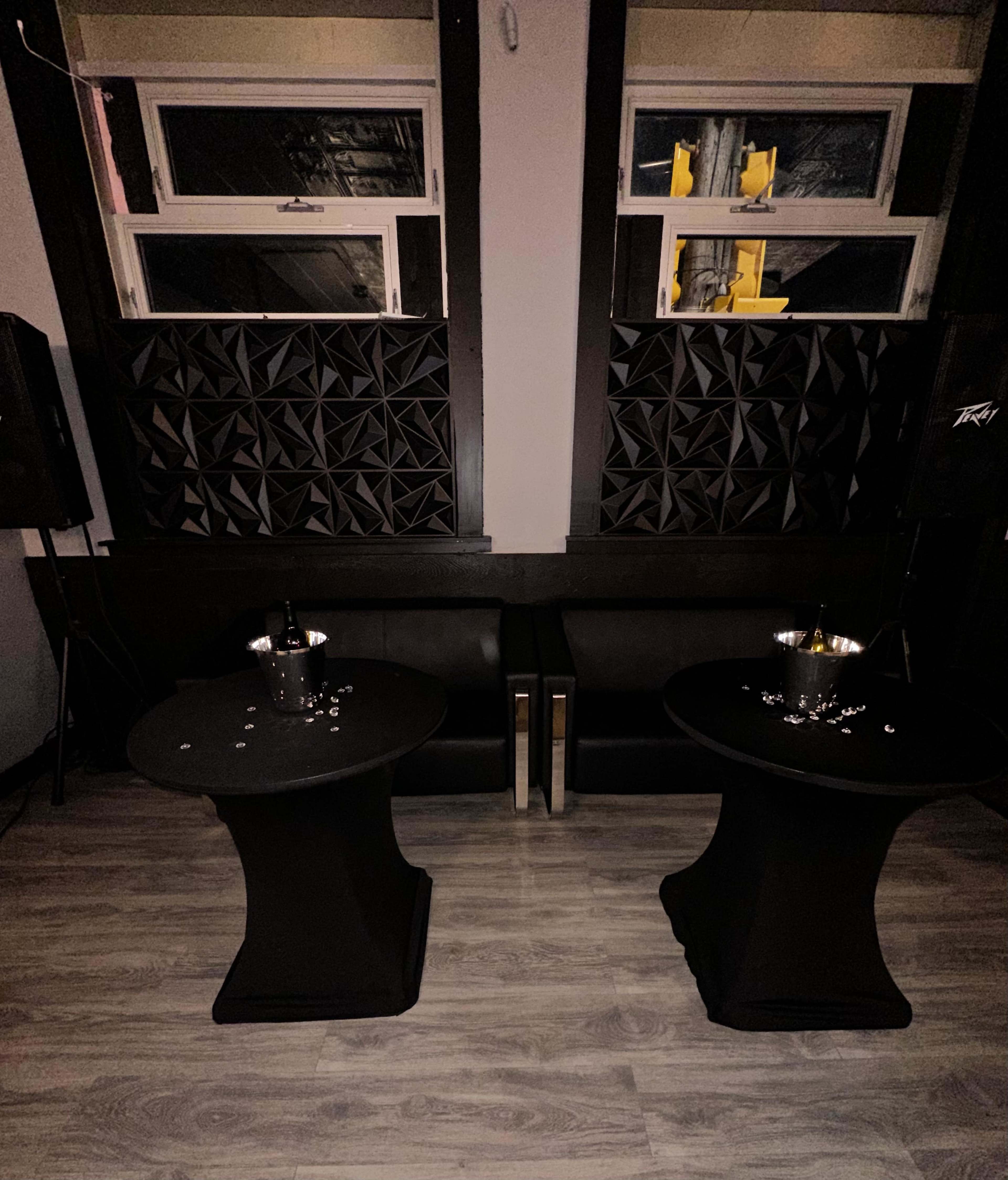 The image shows two black cocktail tables with empty ice buckets on top, positioned in front of a dark sofa in a dimly lit room.