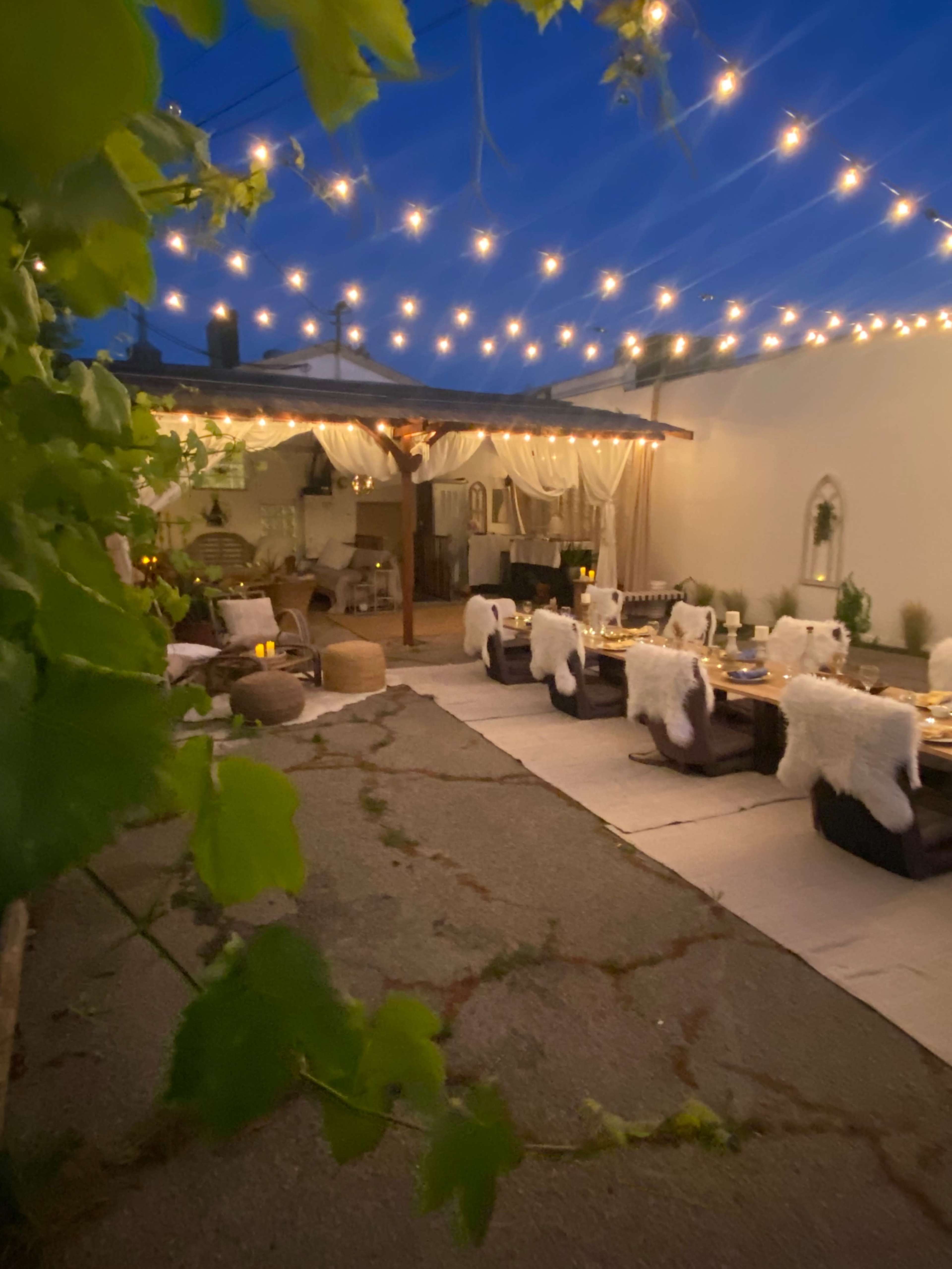 A nighttime outdoor dining area is illuminated by string lights, featuring tables set for a meal amidst lush greenery and cozy seating.
