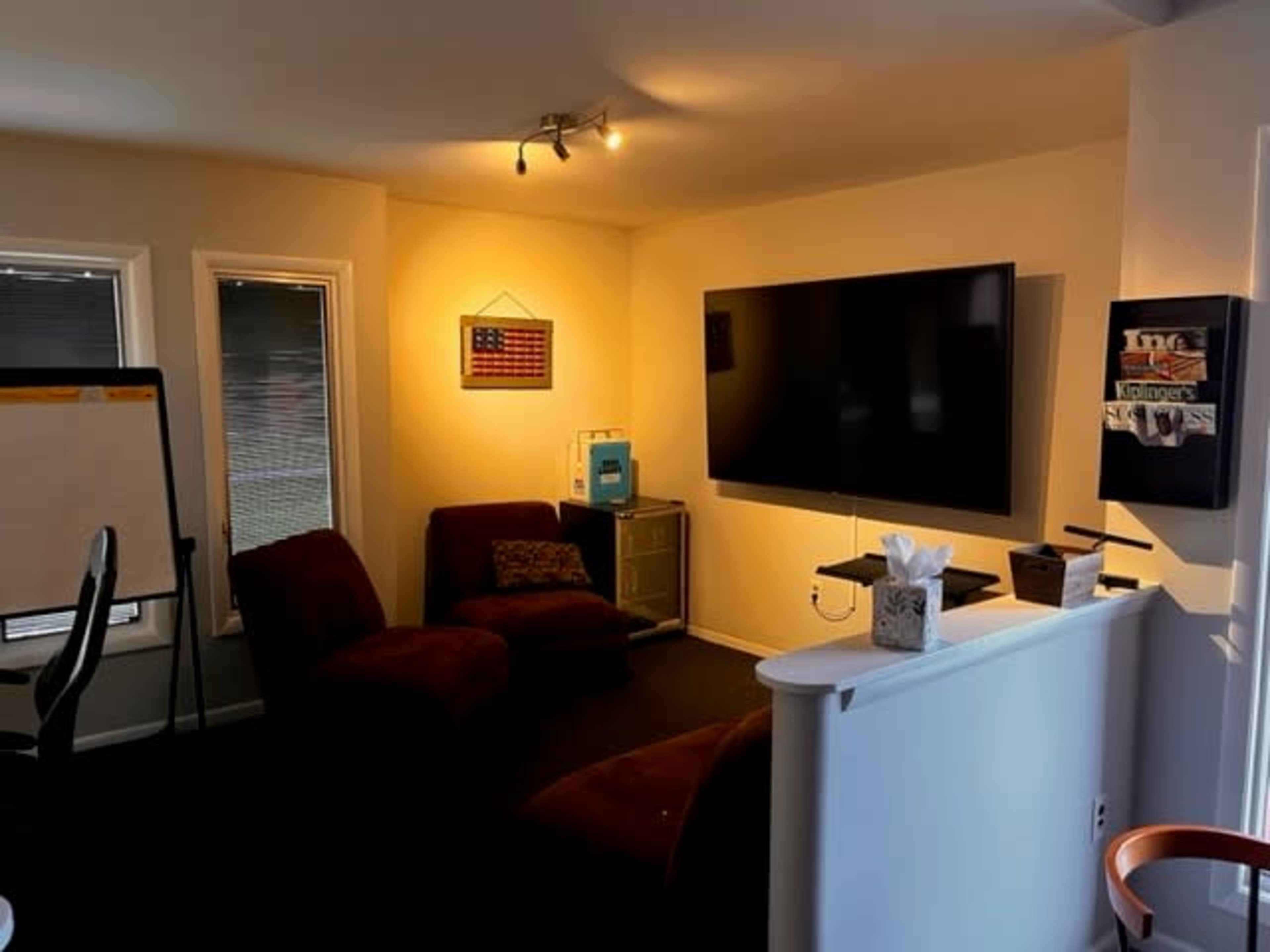 A small room features two brown chairs, a large television mounted on the wall, and a whiteboard beside a window.