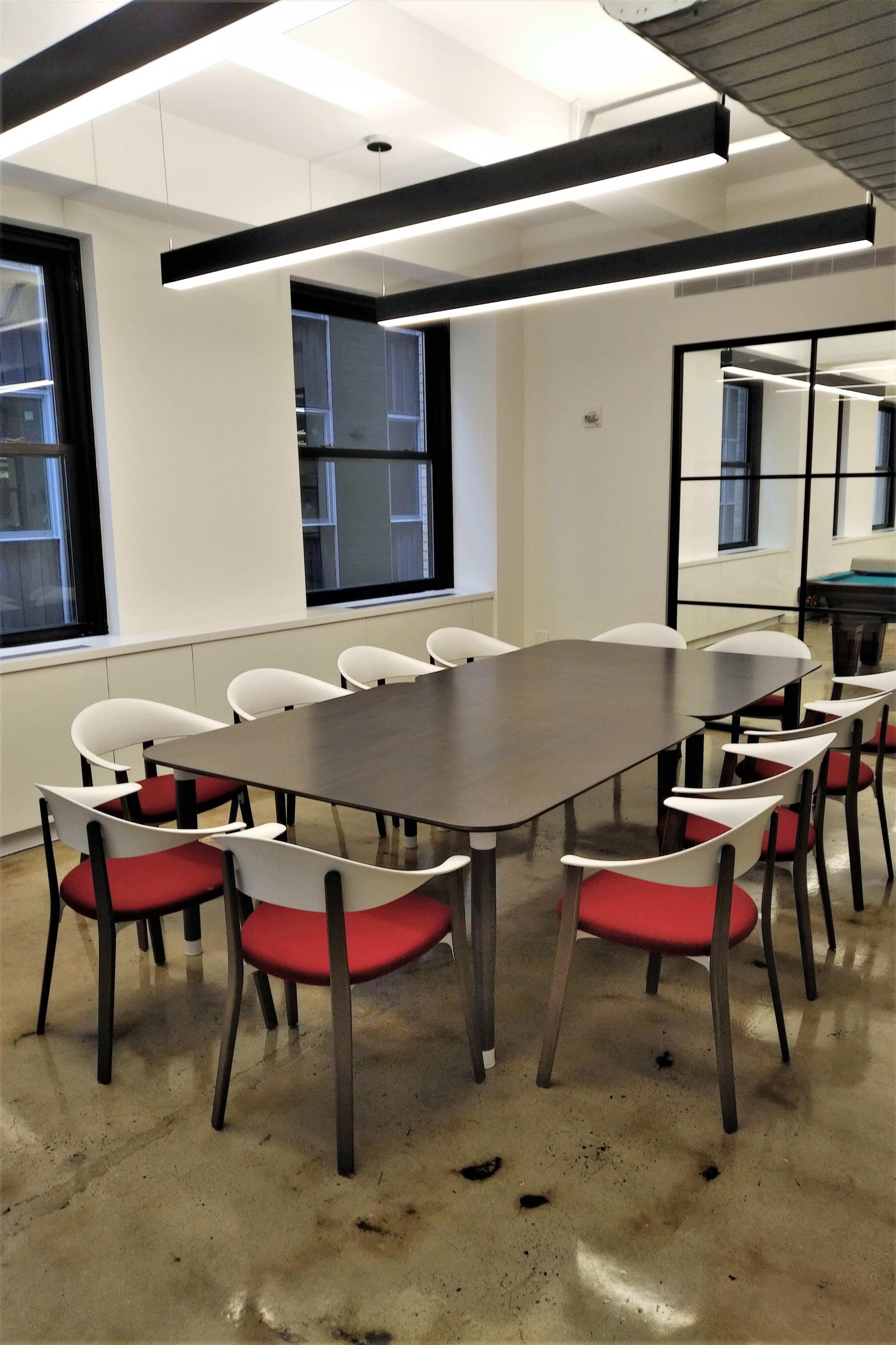A modern conference room features a rectangular wooden table surrounded by eight chairs with white backs and red cushions, set against a backdrop of large windows and a mirror.