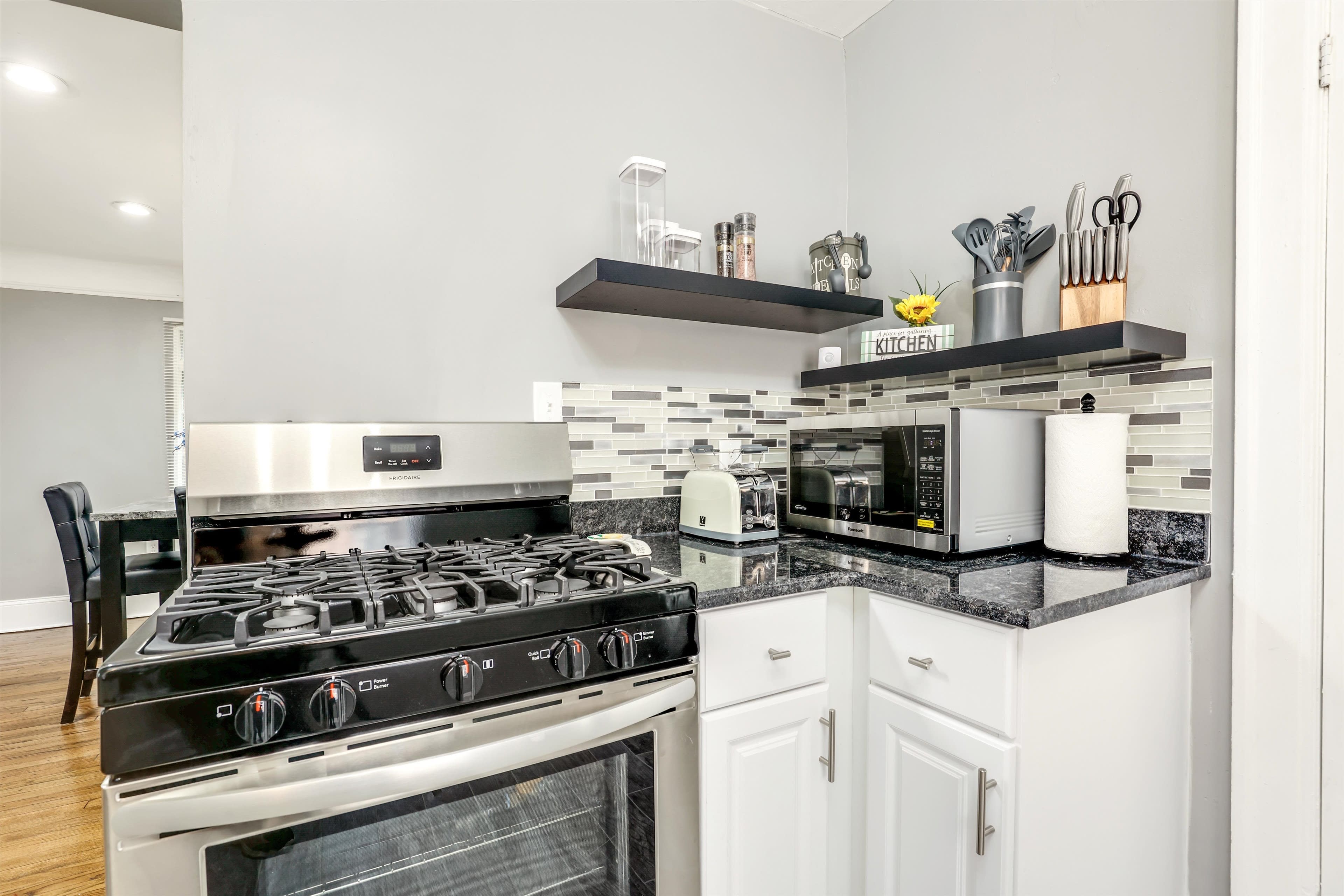 A corner kitchen features a gas stove, stainless steel appliances, and a black granite countertop with decorative tiles and open shelving.