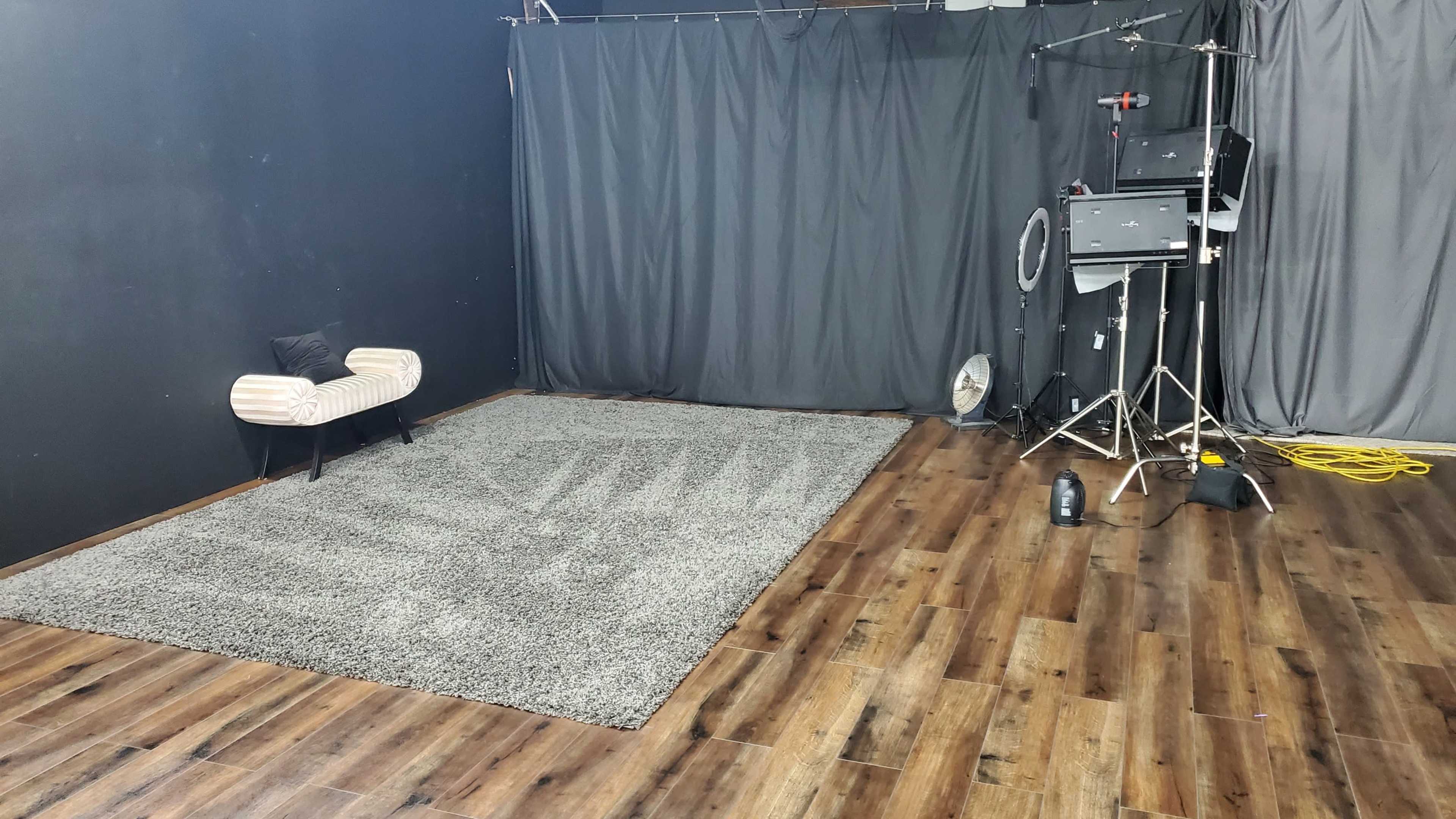 A photography studio with a gray rug, a striped chair, and various lighting equipment set against dark walls and drapes.