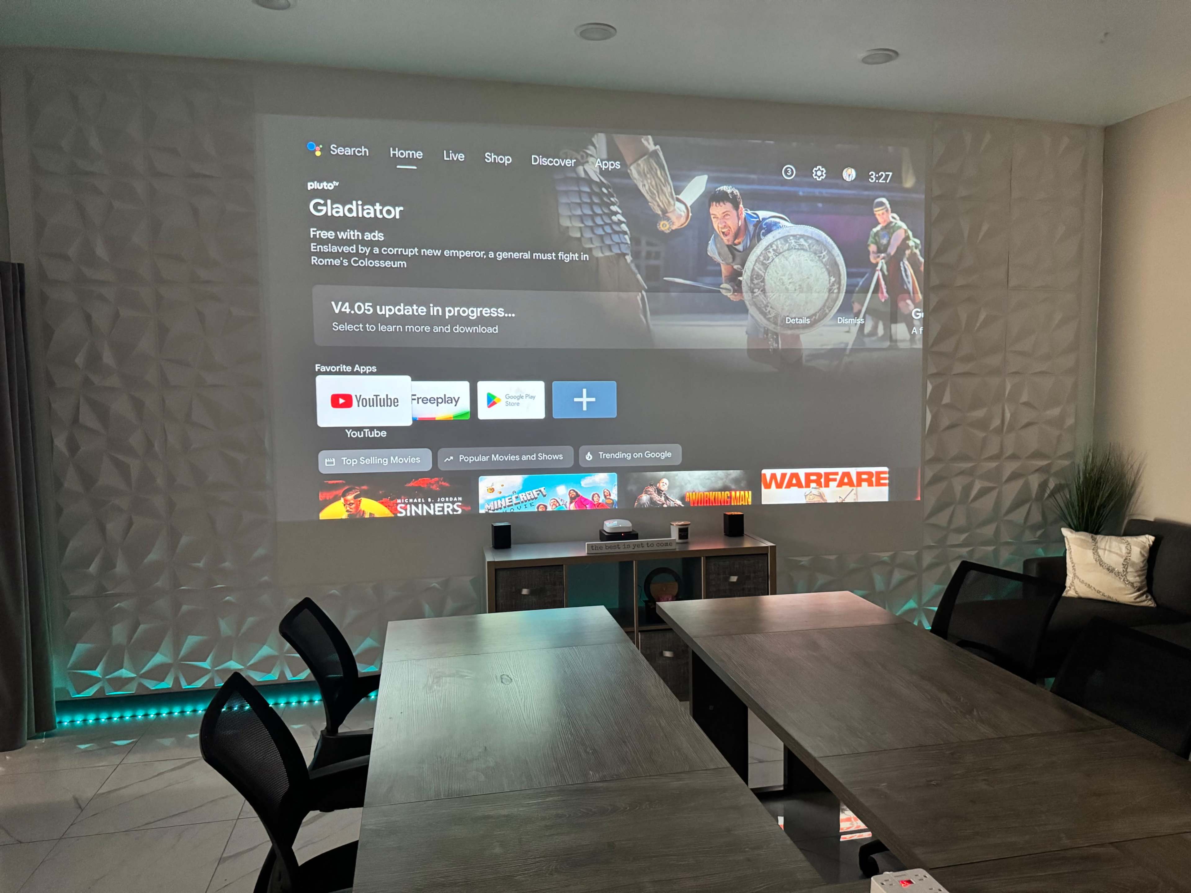 A large projector screen displays a movie selection interface featuring the title "Gladiator" along with icons for apps like YouTube and Freeplay.