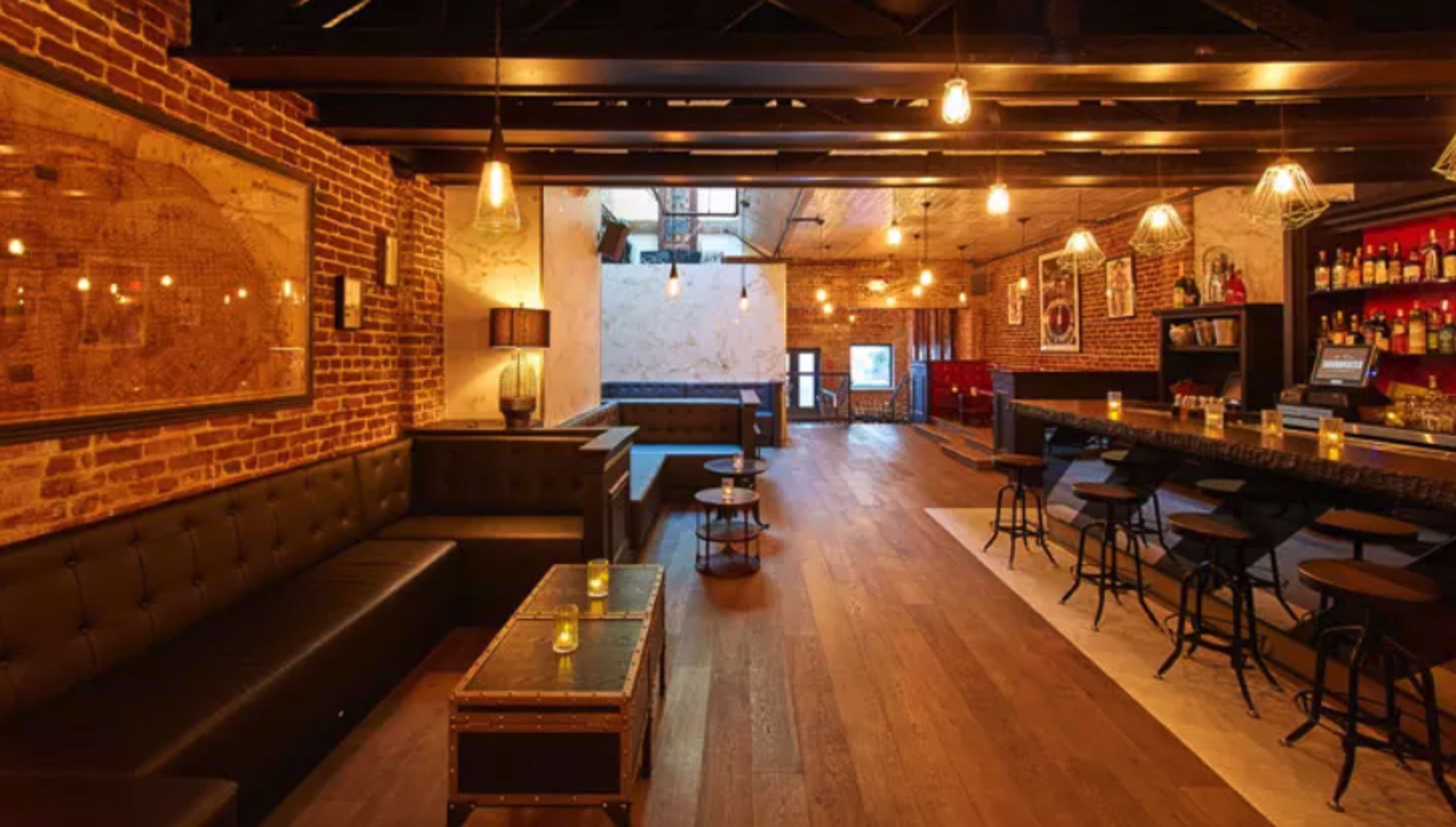 The image shows a contemporary bar interior with exposed brick walls, wooden flooring, and a long bar topped with various bottles and illuminated by pendant lights.