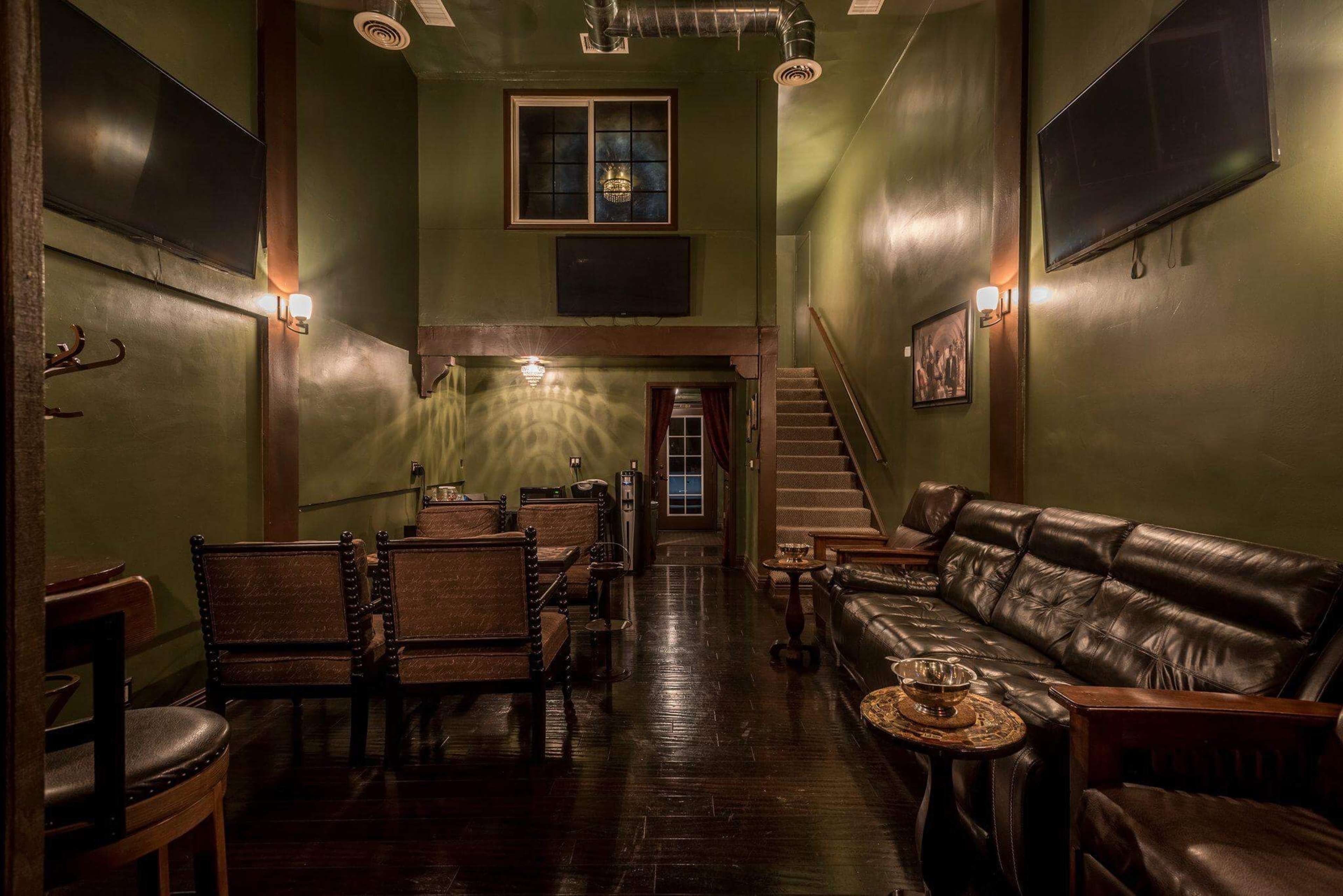 A dimly lit lounge with green walls, black wooden floors, leather seating, and a staircase leading to a second level.