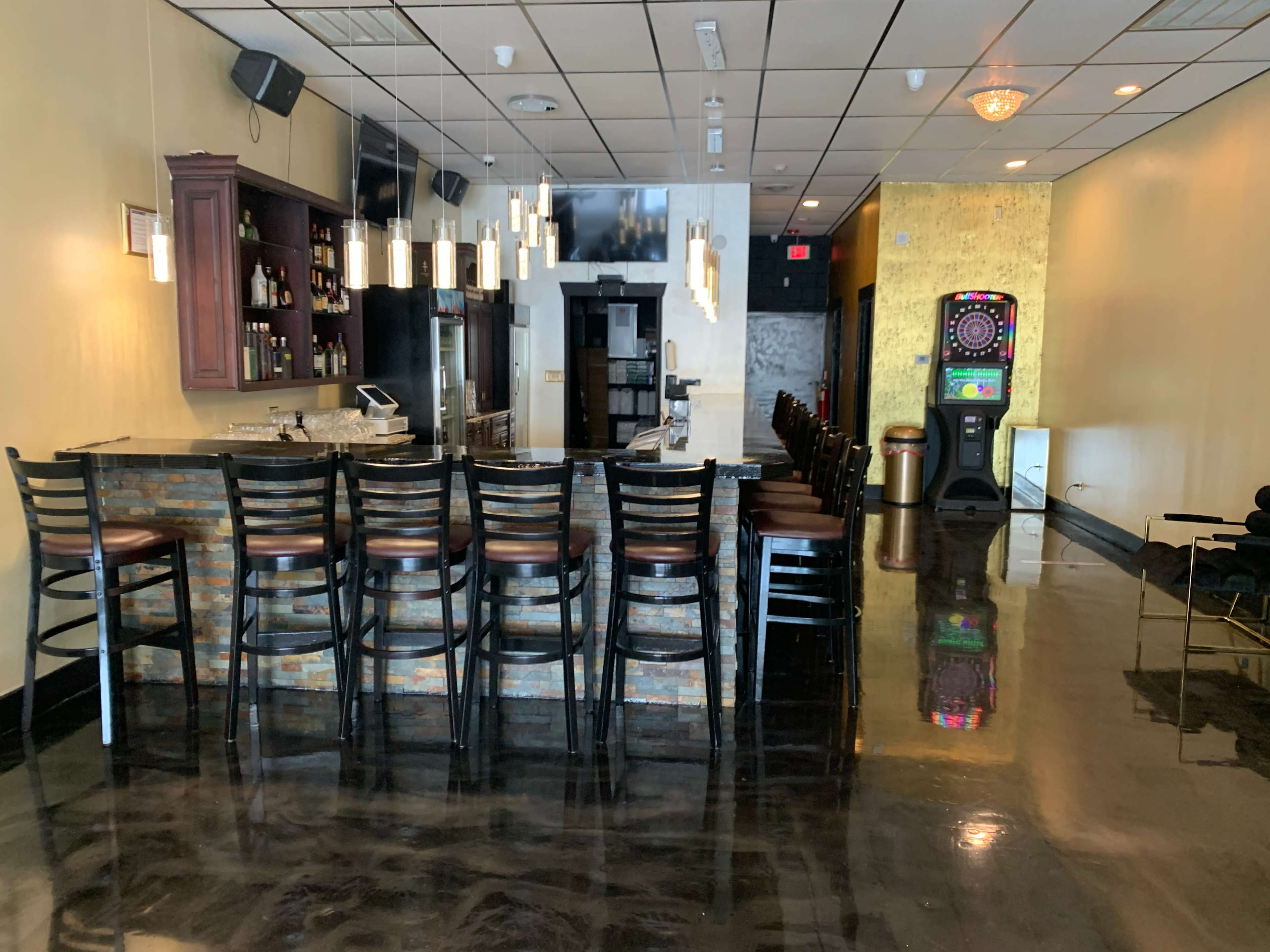 An empty bar area with a polished black floor, wooden stools lined up at the counter, and a dartboard machine in the corner.