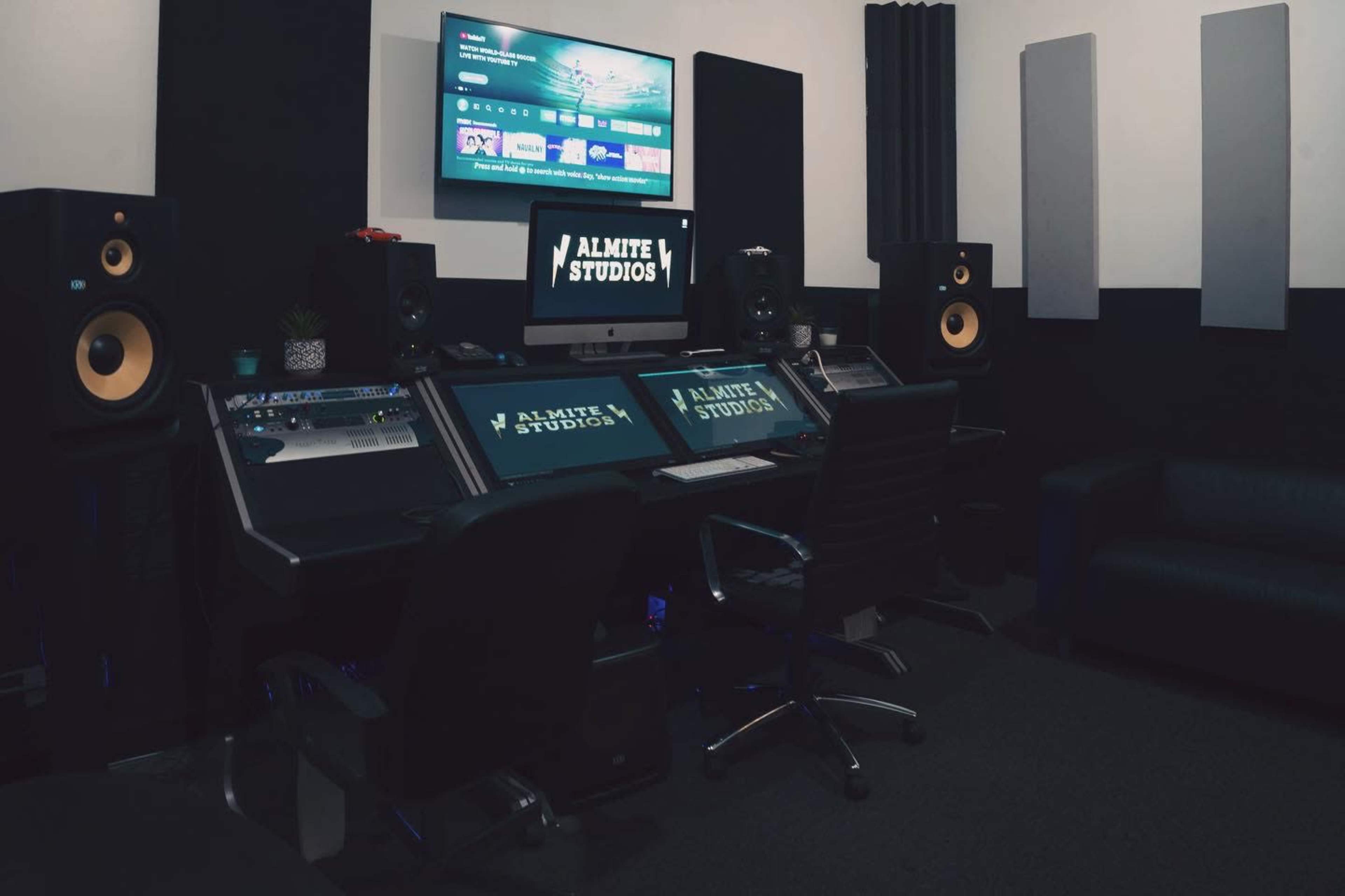 A music studio equipped with a digital mixing console, speakers, a television screen, and soundproofing panels on the walls.