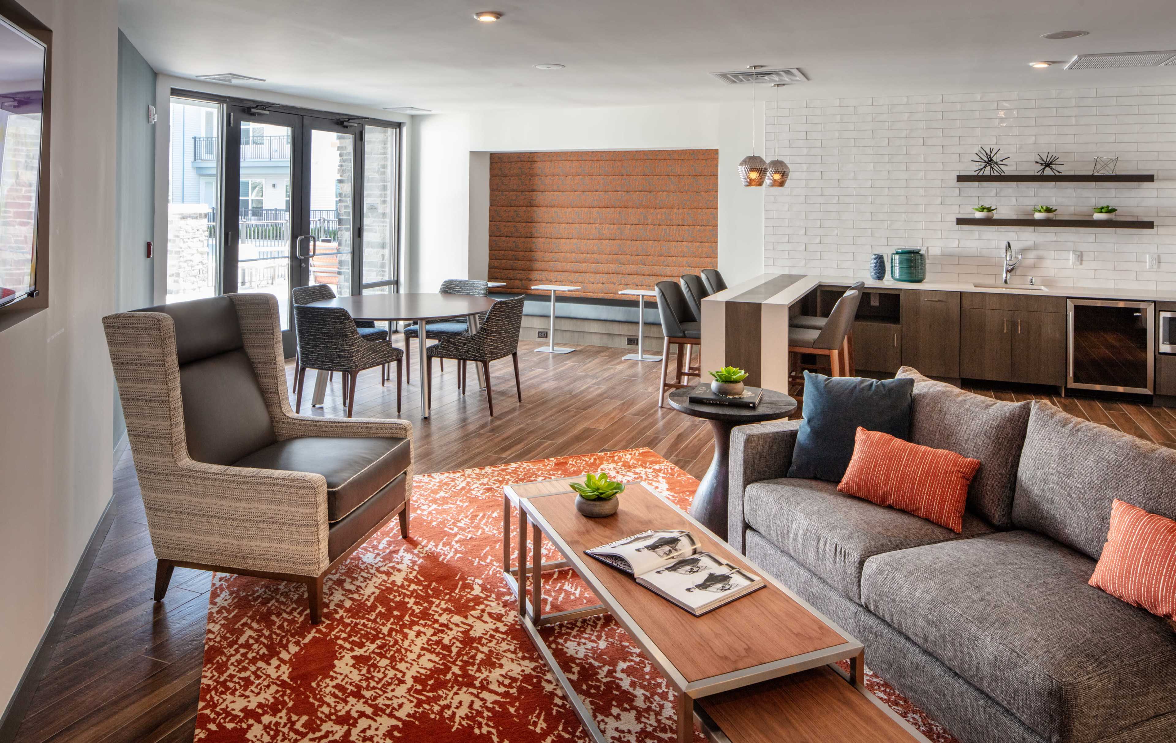 A modern living area with a gray sofa, orange accent pillows, a coffee table, and a dining space featuring a round table and chairs, all set in a well-lit, open-layout room with large windows.