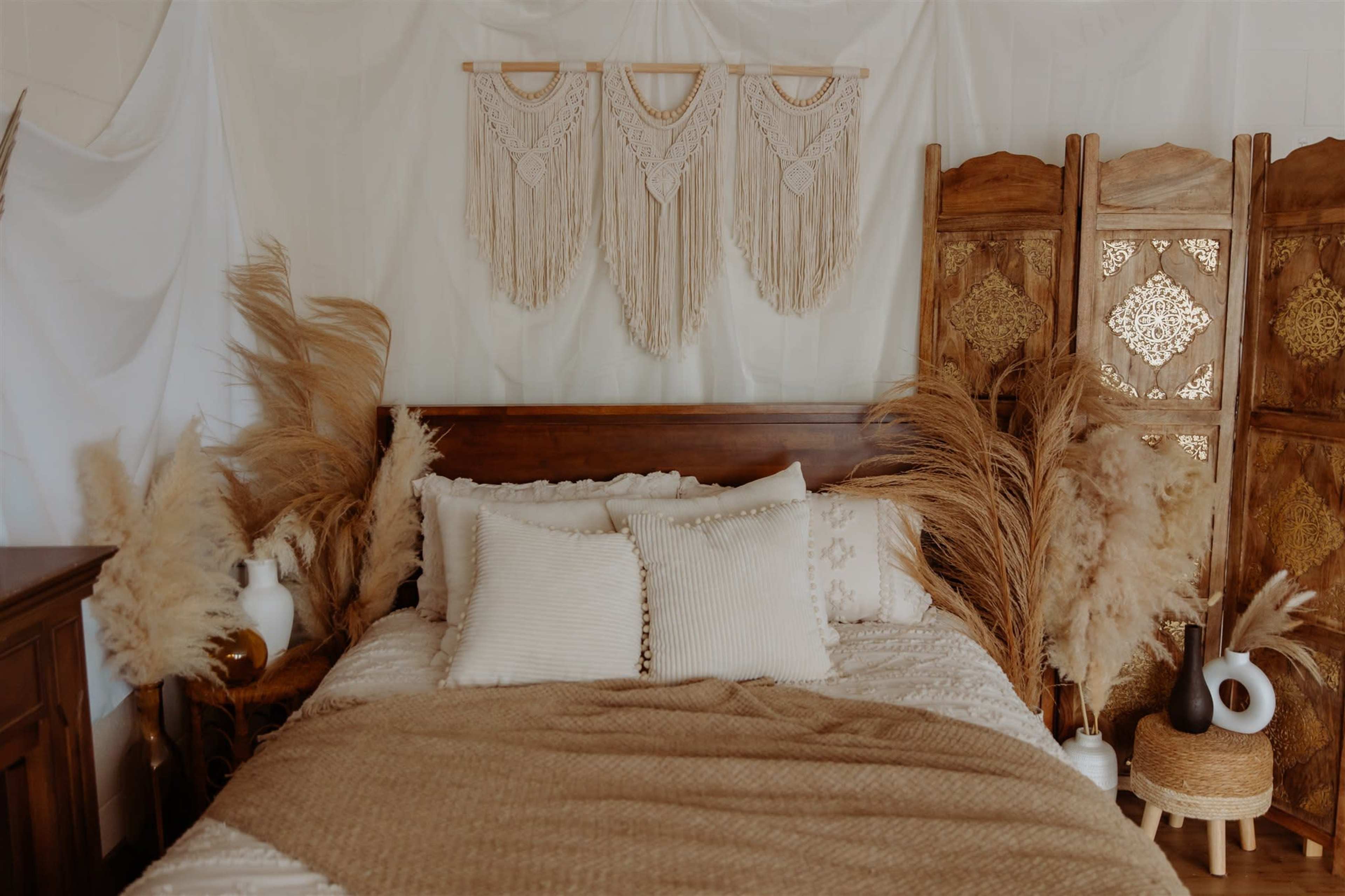 A wooden bed with decorative pillows is surrounded by pampas grass and a folding screen, against a backdrop of draped fabric and macramé wall hangings.