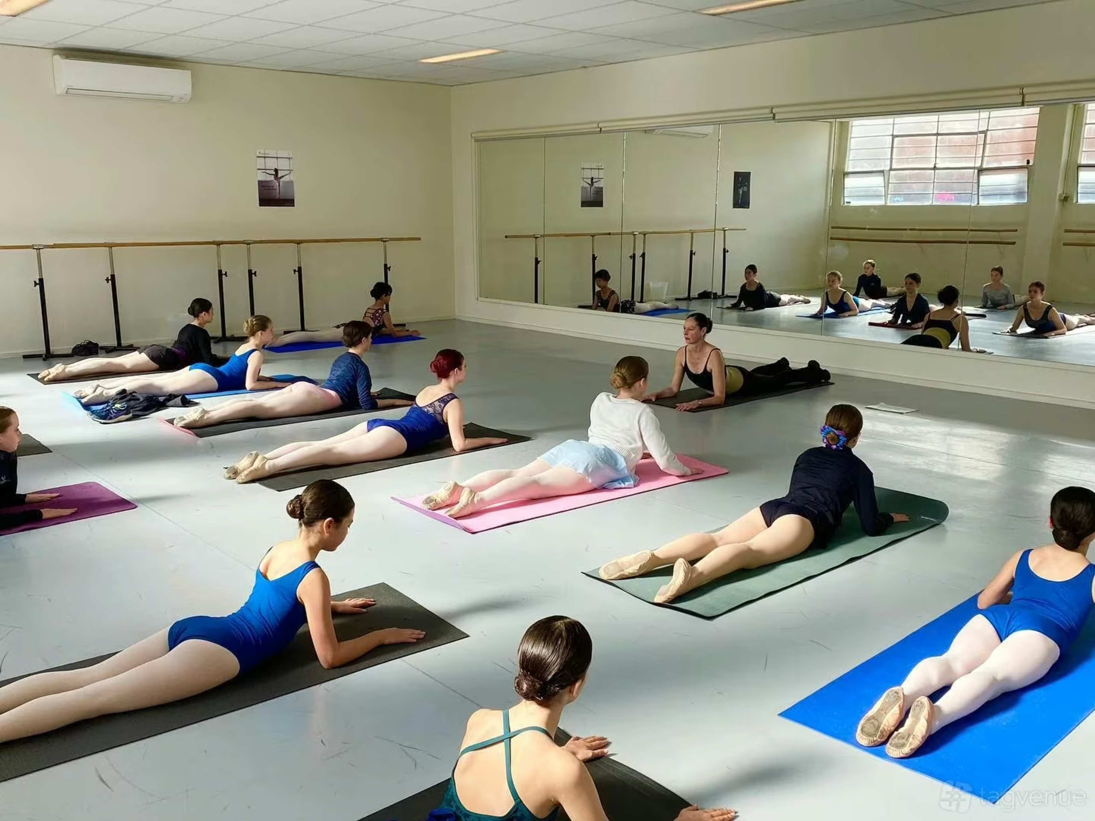 A ballet class is taking place in a studio, with students performing exercises on mats while mirrored reflections show their positions.