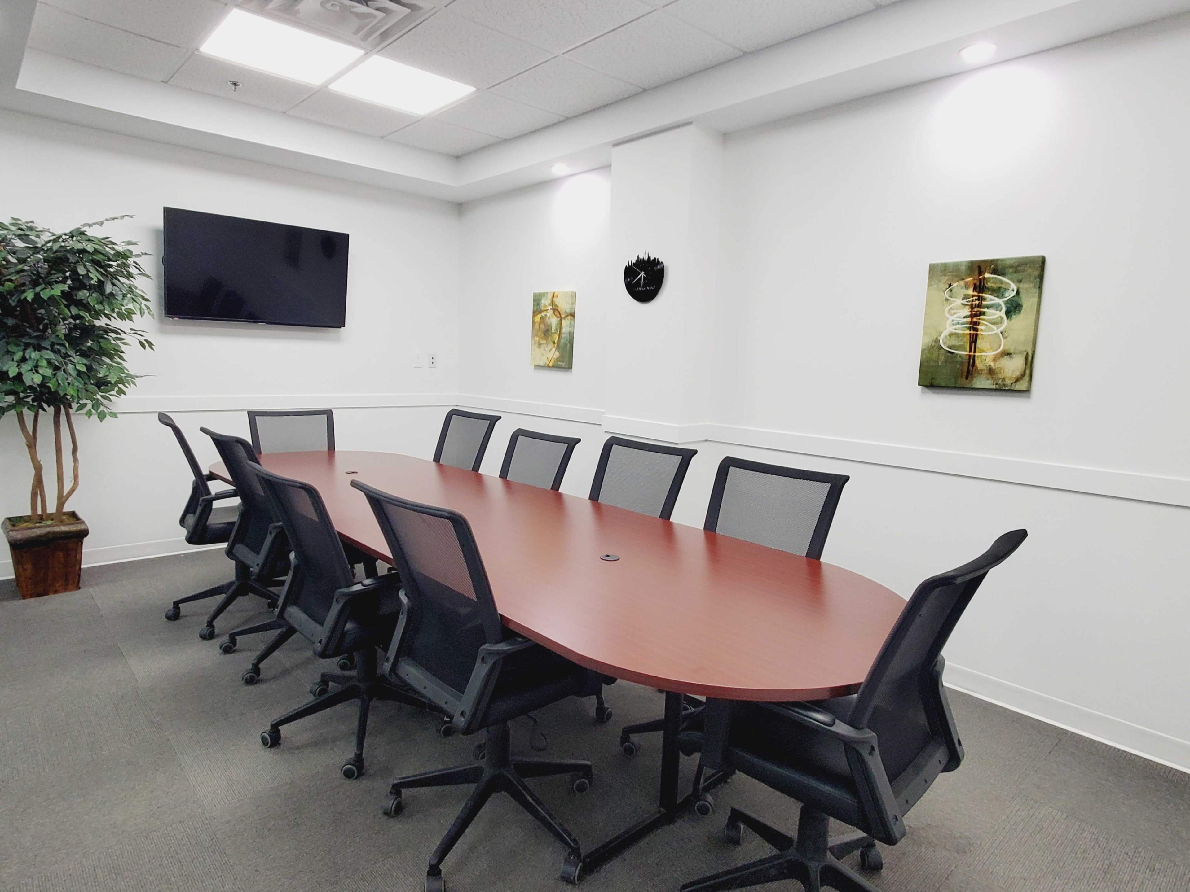 A conference room features an oval wooden table surrounded by black chairs, with a TV and two framed artworks on the walls.