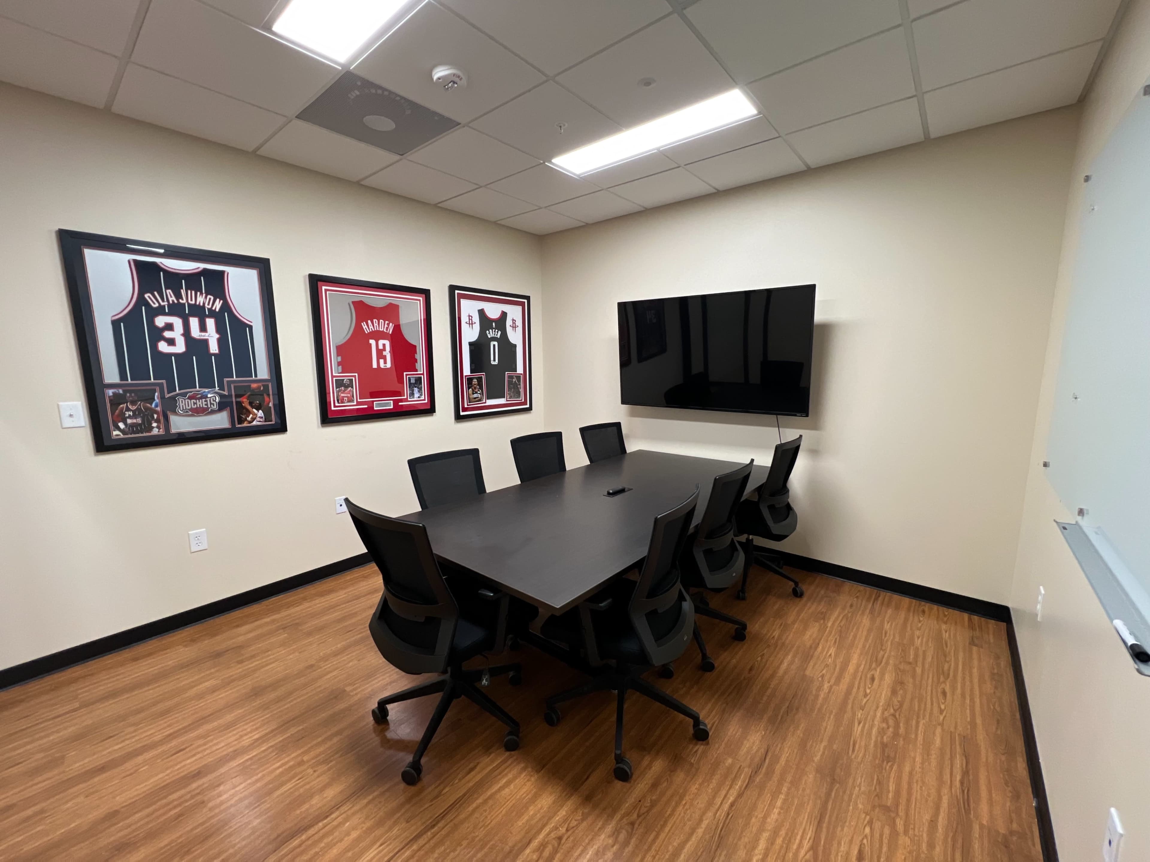 A conference room features a long black table surrounded by chairs, with sports jerseys displayed on the walls and a wall-mounted television.