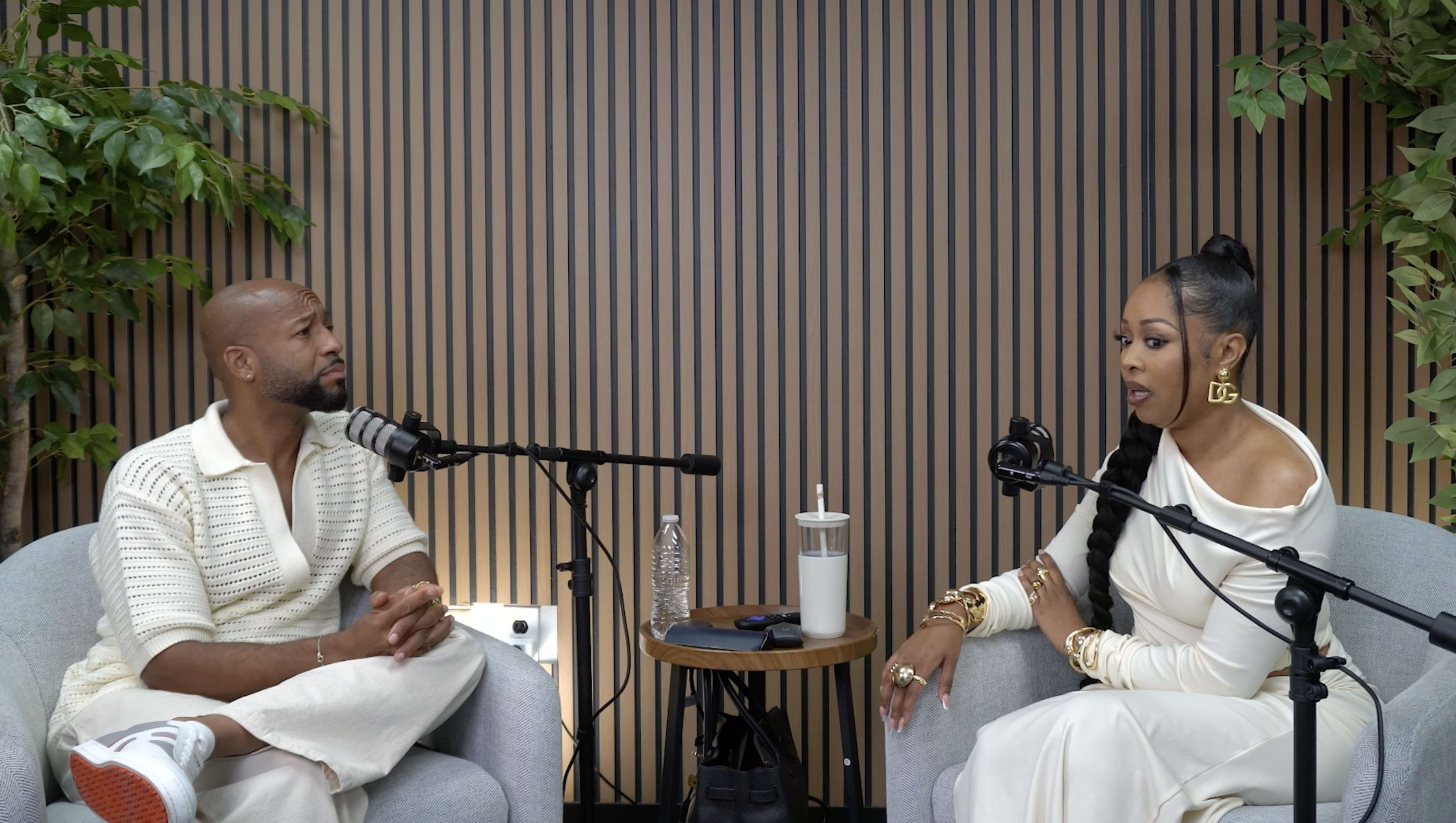 Two individuals are seated in a modern, minimalistic setting with microphones, engaging in conversation, while the backdrop features vertical wooden panels and some greenery.