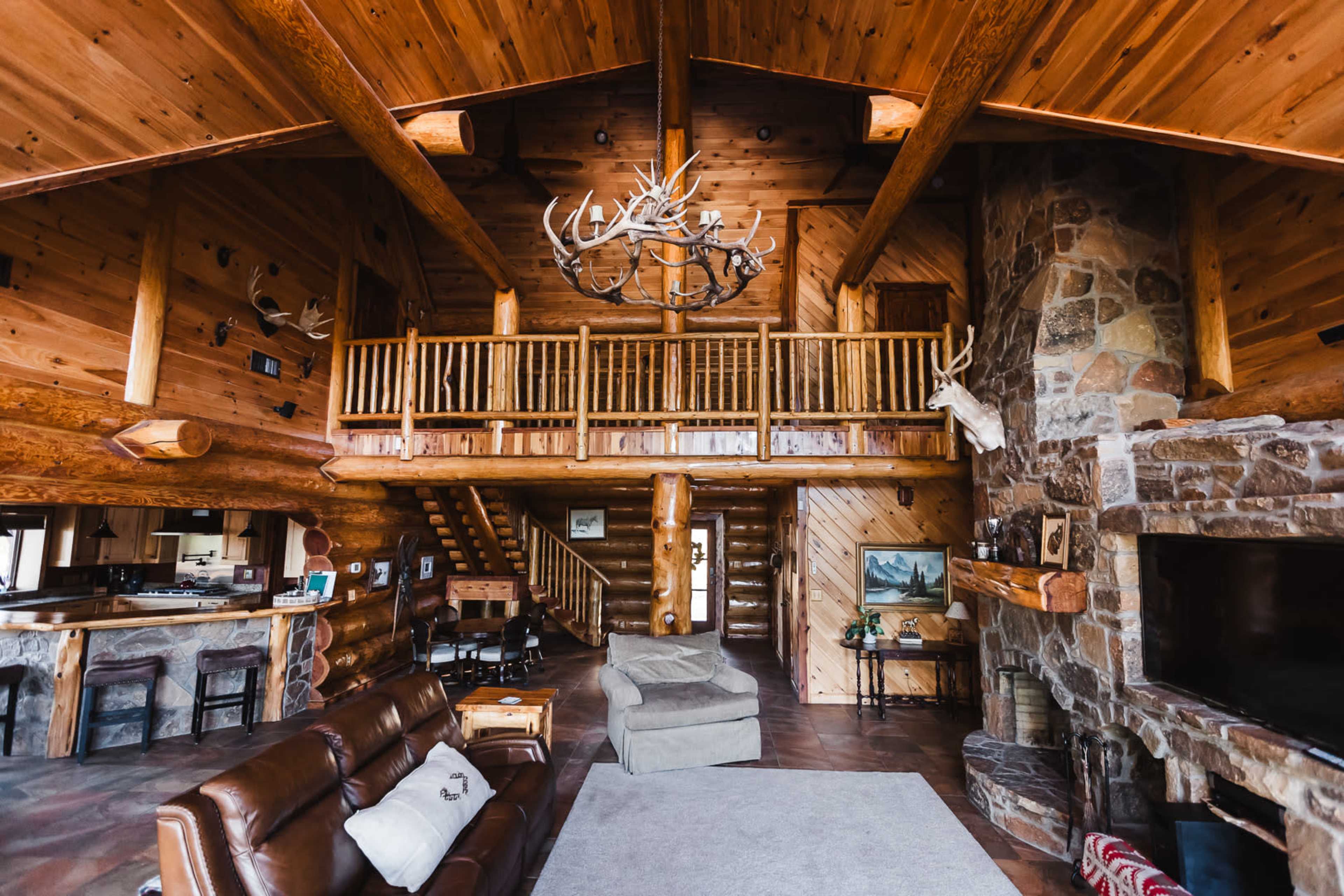 The image shows a spacious log cabin interior with high ceilings, wooden beams, a stone fireplace, and a second-floor balcony.