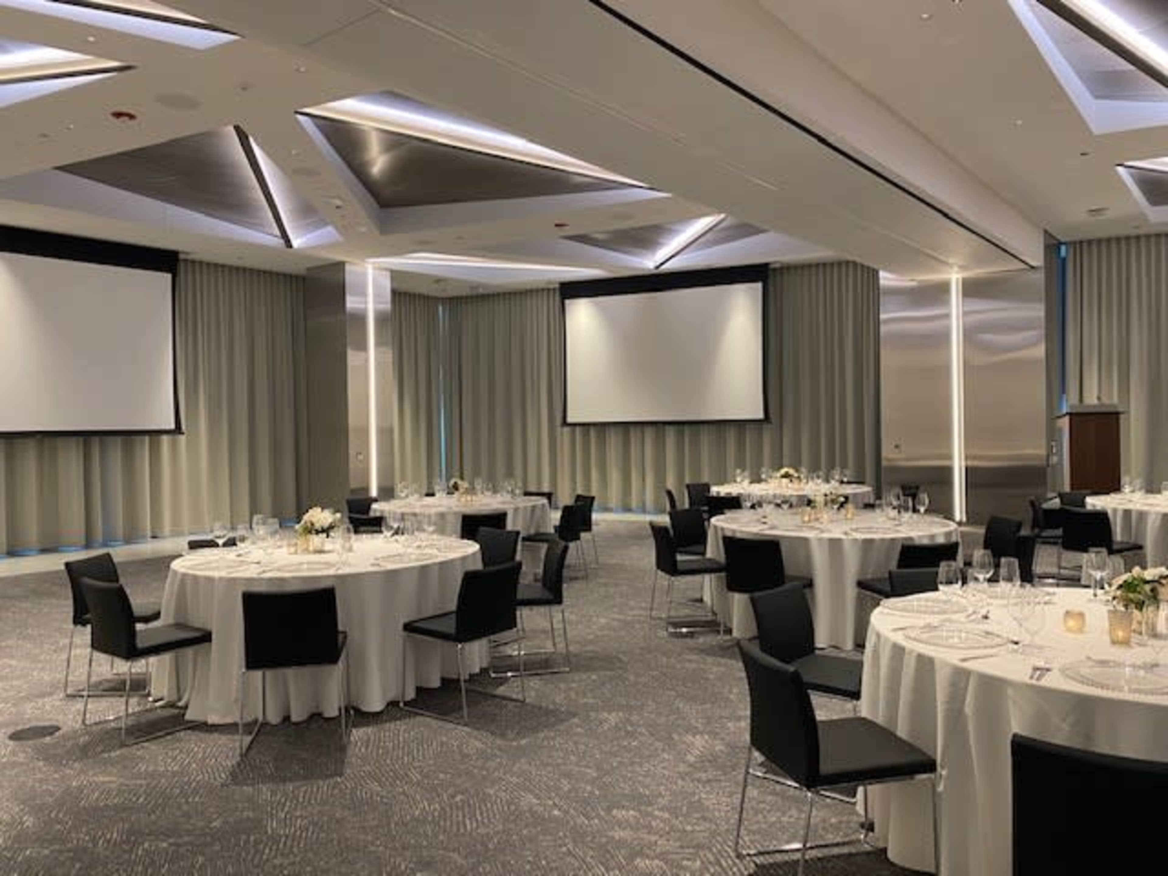 A banquet hall is set up with round tables covered in white tablecloths, surrounded by black chairs, and features screens at the front for presentations.