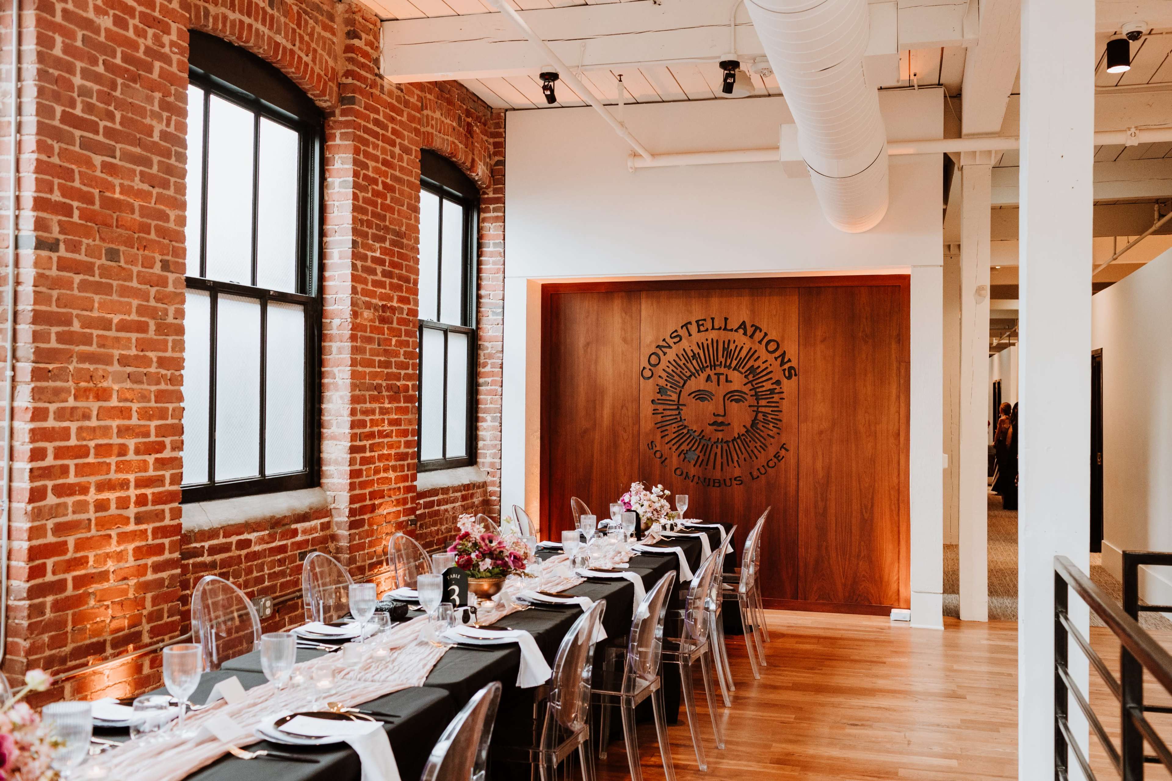 The image shows a well-decorated dining area with a long table set for a meal, featuring exposed brick walls and a wooden panel displaying a sun emblem.