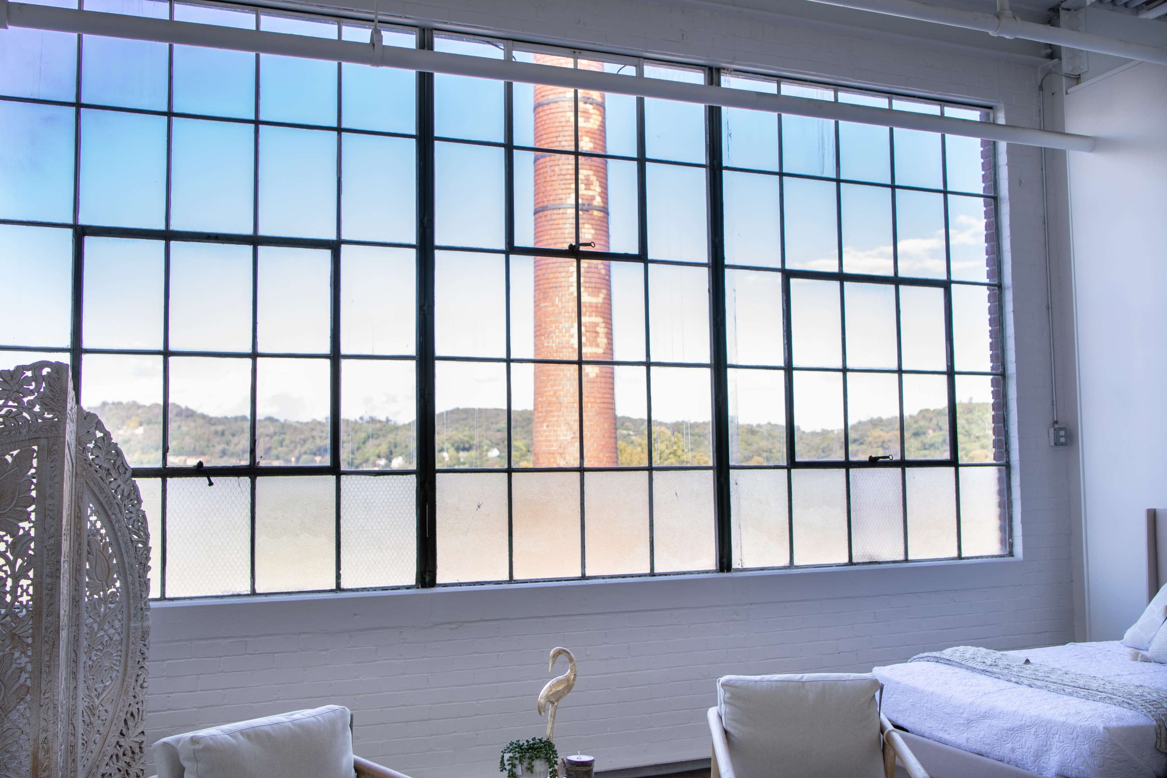 A large industrial window with a brick chimney visible outside, framed by white walls and a simple interior setup.