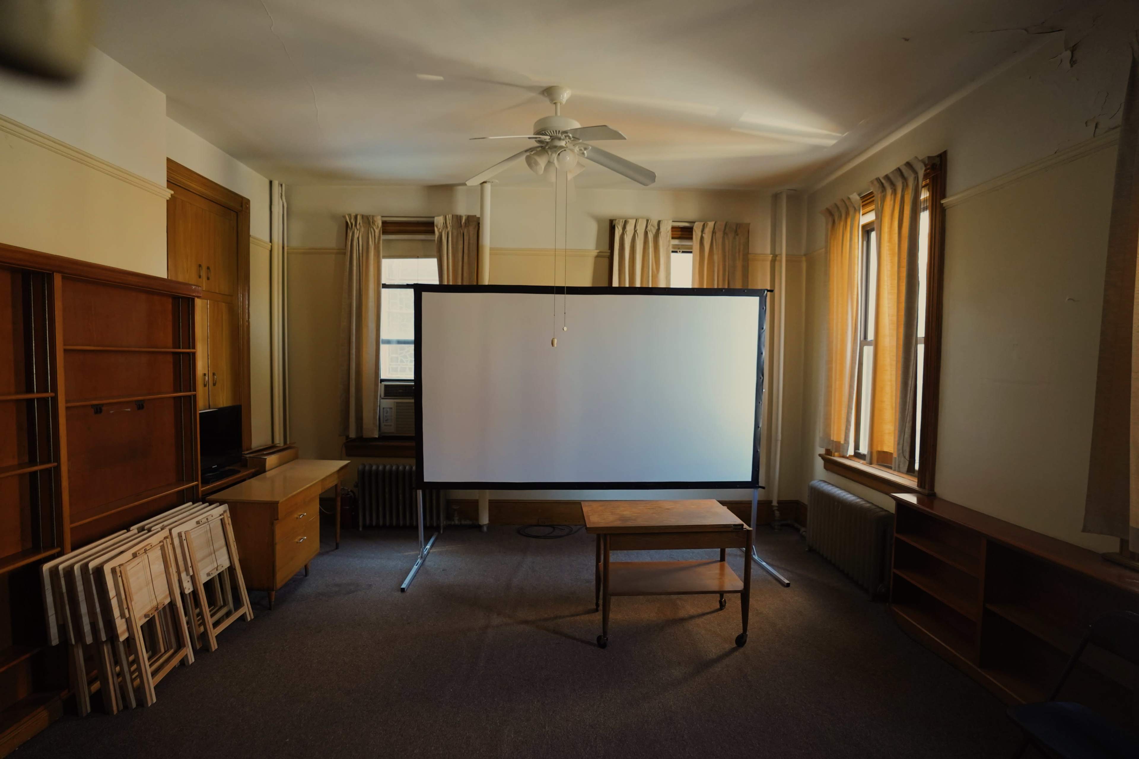 An empty room with a large projection screen at the center, surrounded by wooden furniture and windows with white curtains.