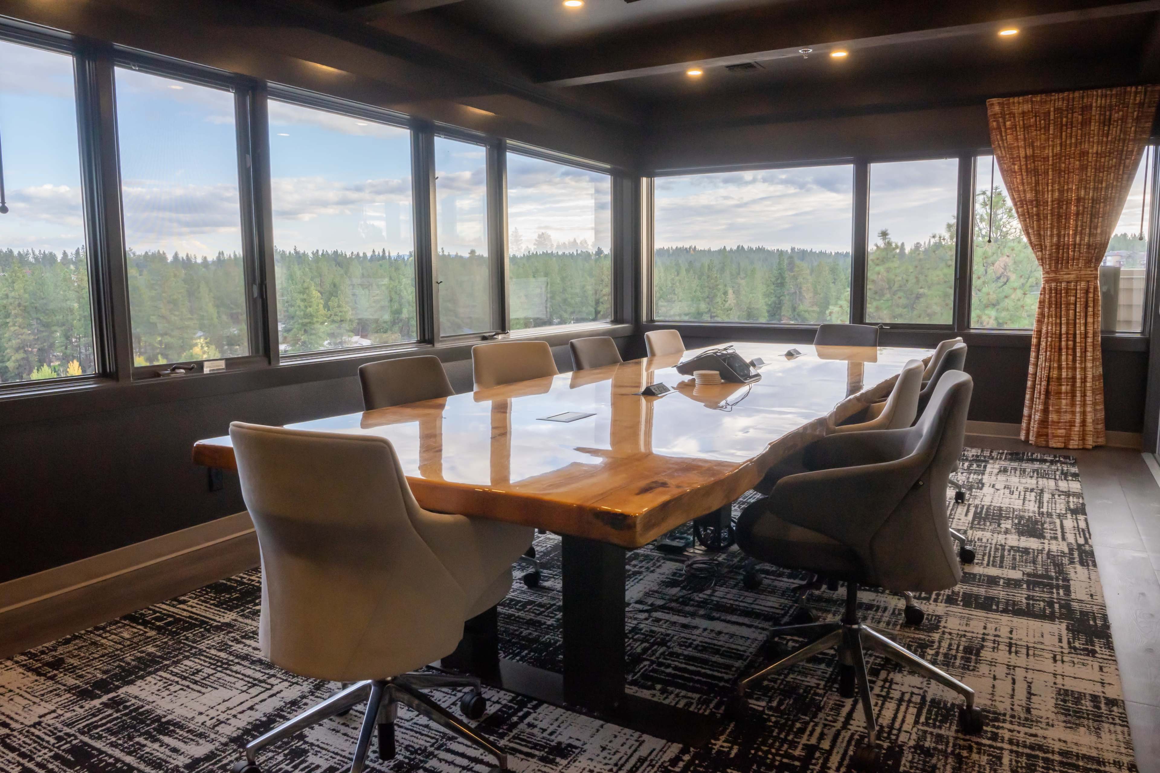 A conference room with a large wooden table surrounded by stylish chairs and large windows overlooking a forested landscape.
