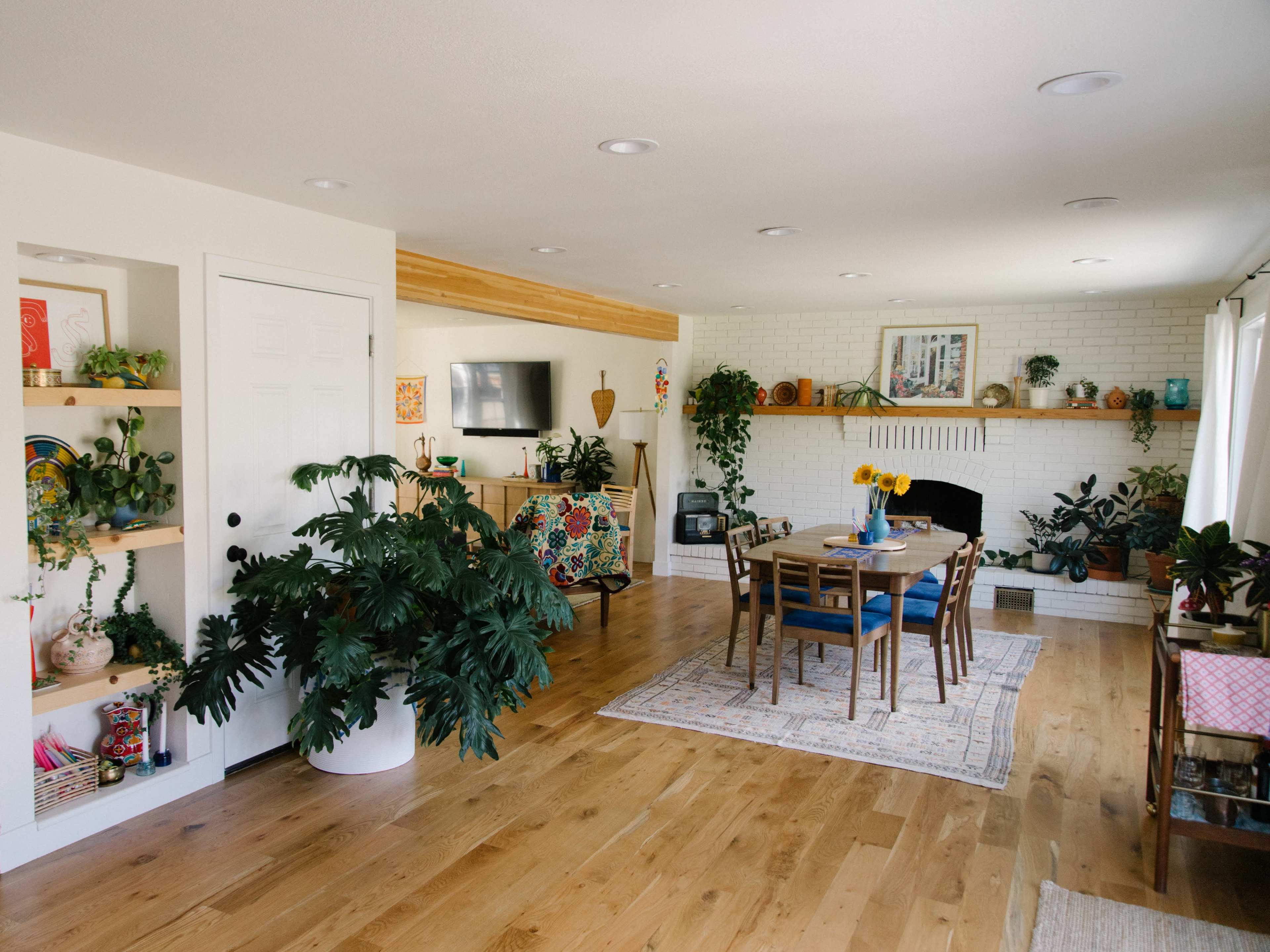 A bright living space features a dining table centered under overhead lights, surrounded by plants, a television, and a cozy fireplace with a light-colored brick surround.