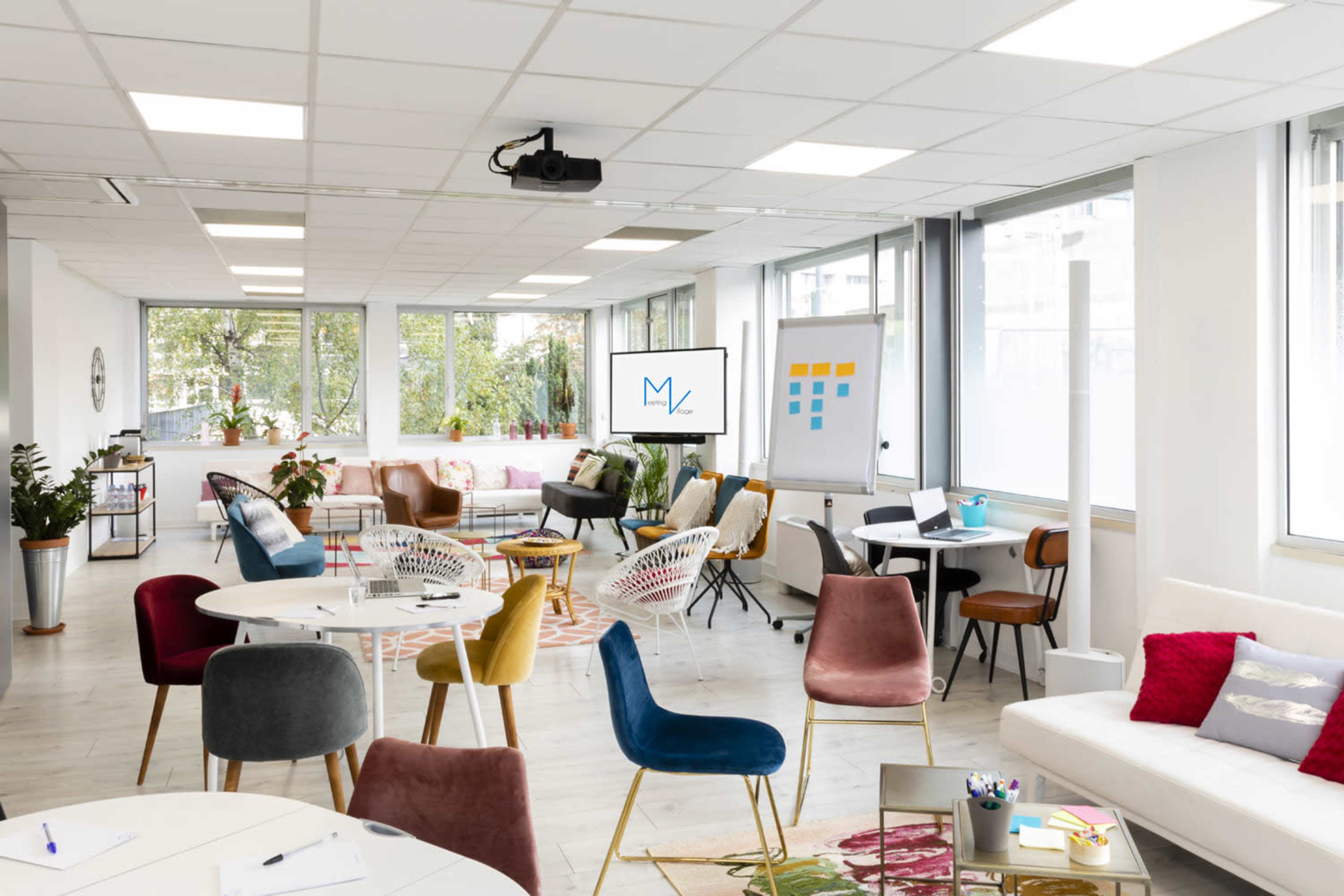 A modern, open office space with various seating arrangements, a projector screen displaying a logo, and large windows allowing natural light to enter.
