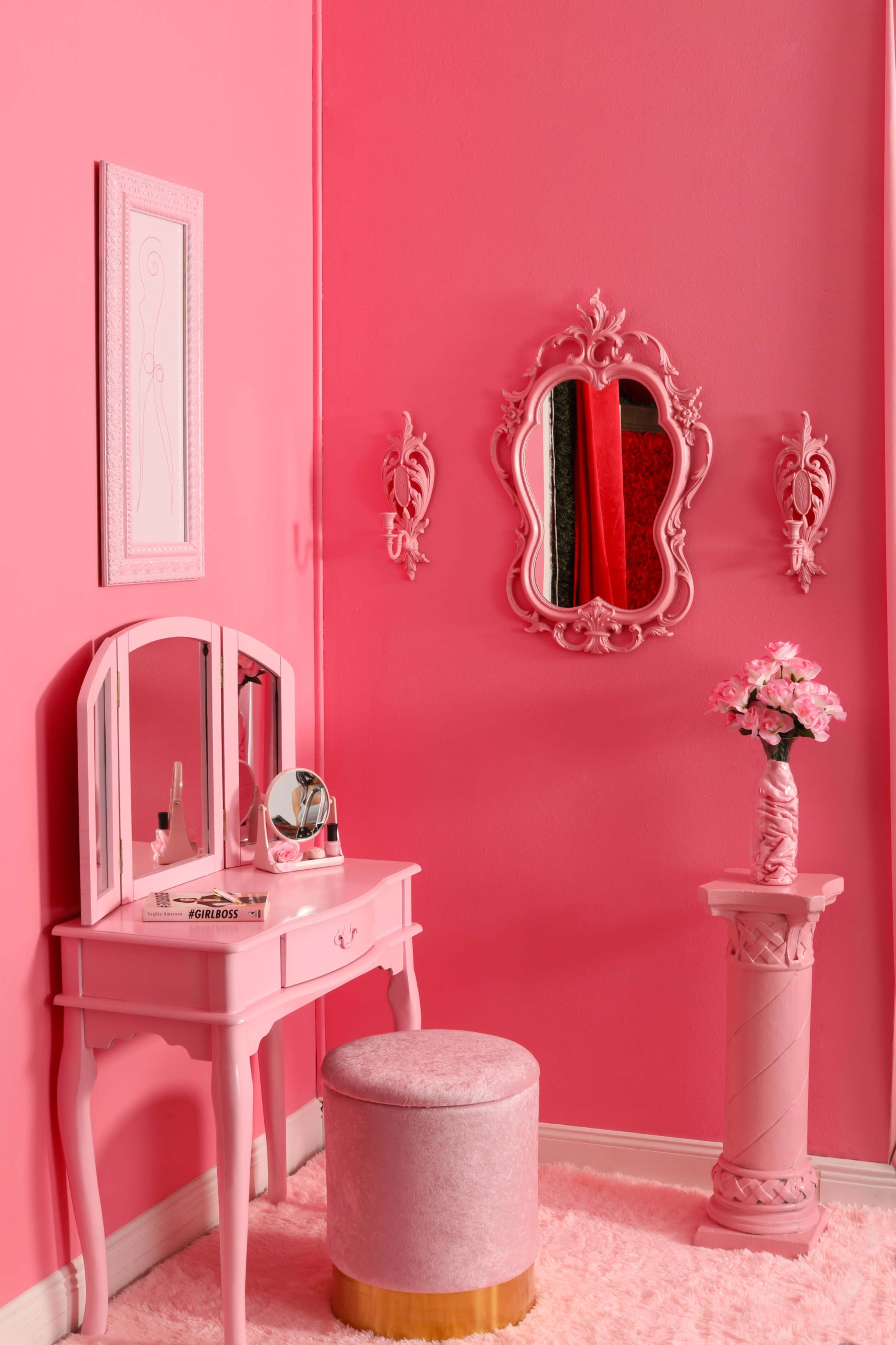 A fully pink room with a vanity table, a round stool, a decorative mirror, and a flower vase on a pedestal.