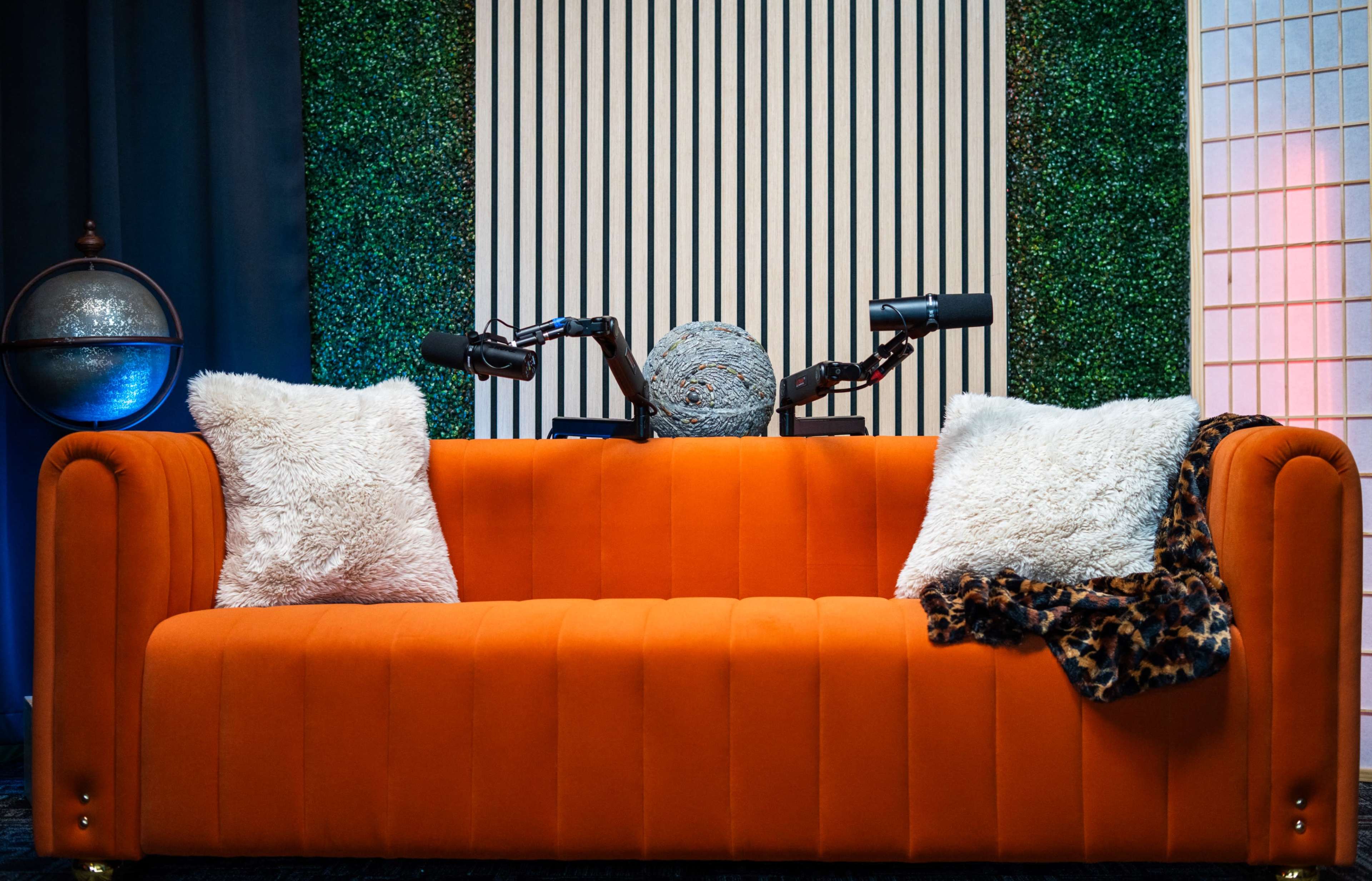 An orange couch with white and leopard print pillows is positioned against a backdrop of green foliage and vertical striped panels, with a scooter resting on the back.