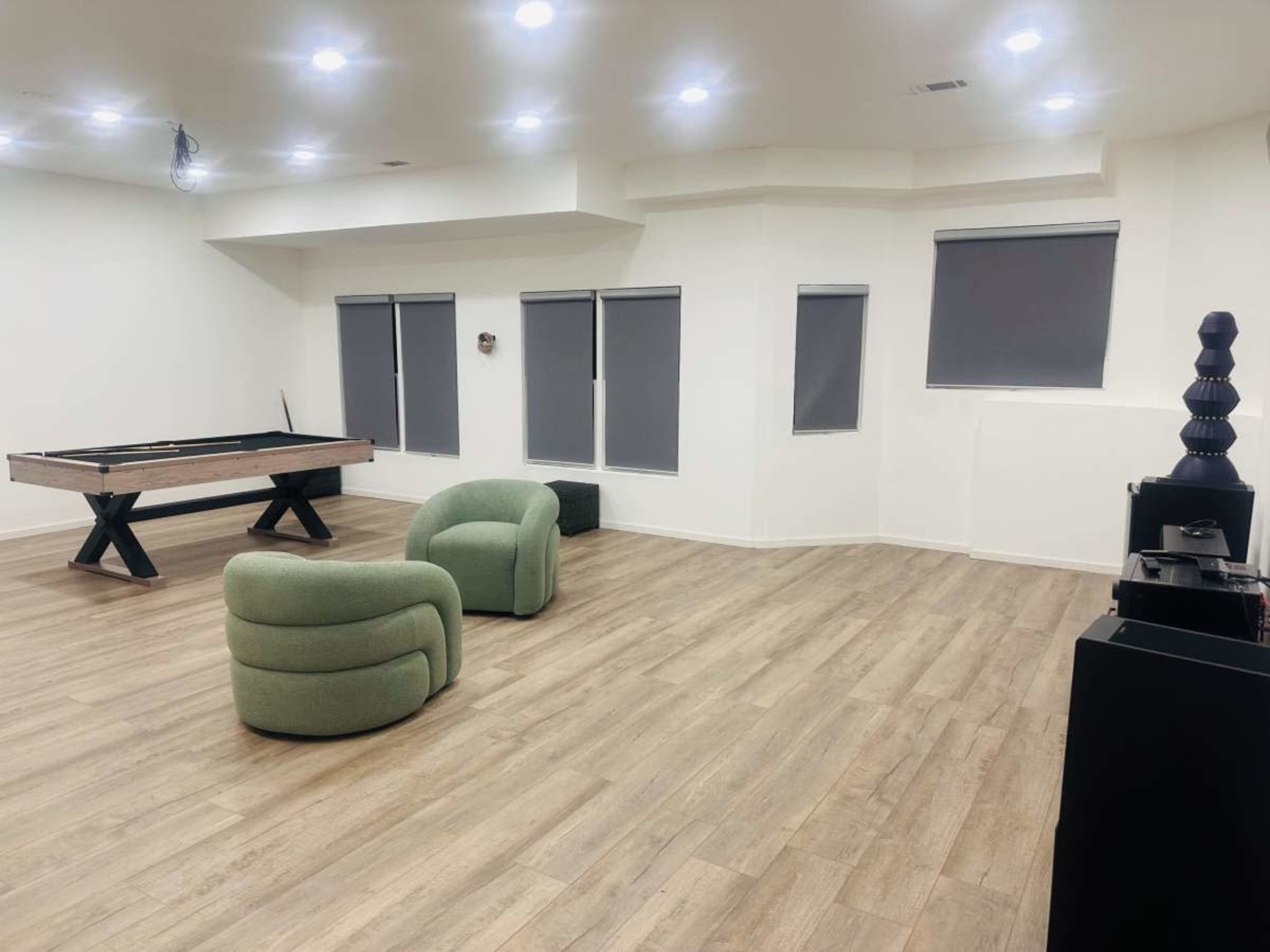 The image shows a spacious, well-lit room featuring a billiard table, two green armchairs, and several windows with dark blinds.