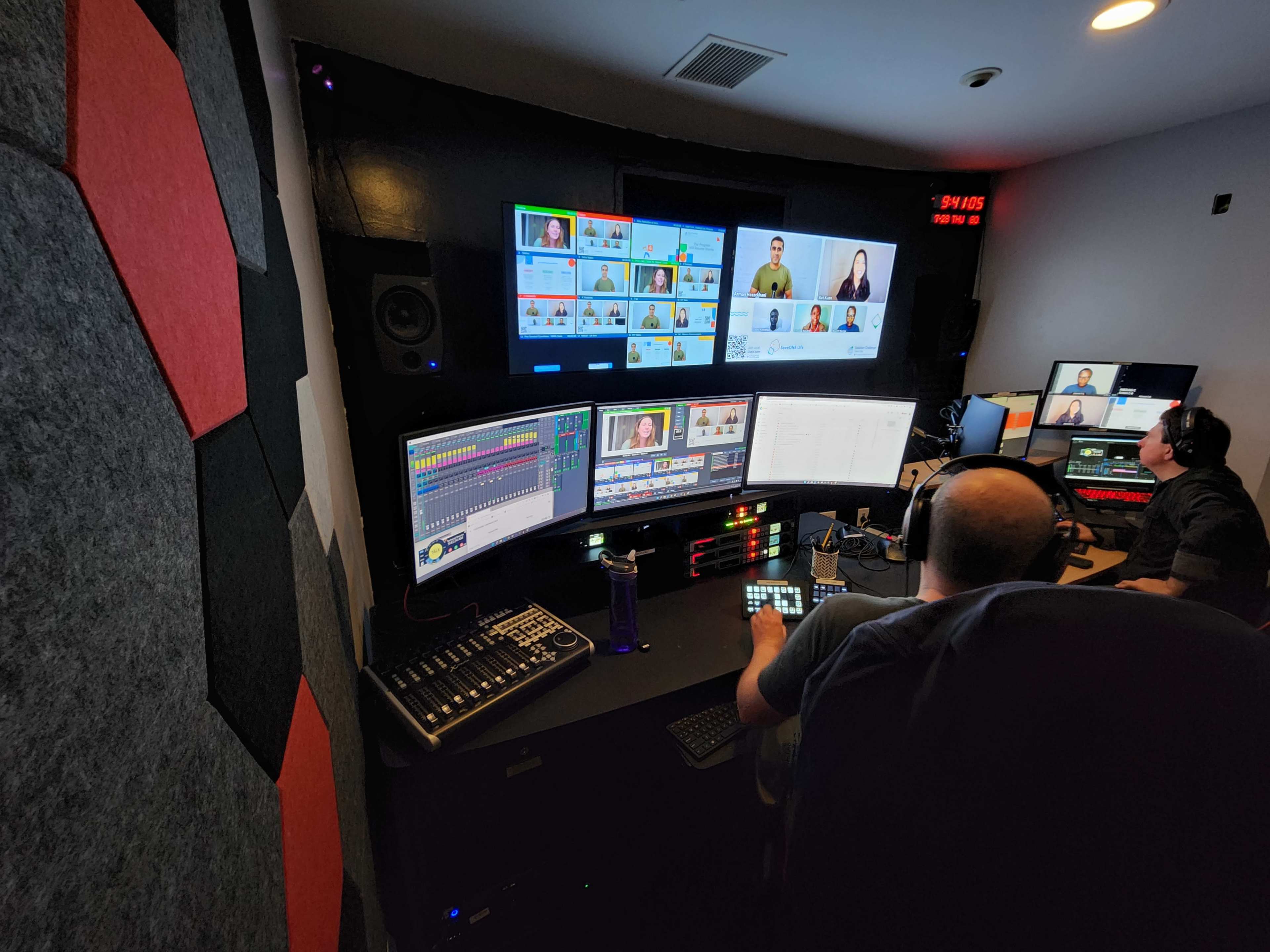 A control room with several large monitors displaying video feeds of people, while two operators are working at computer stations with audio and video equipment.