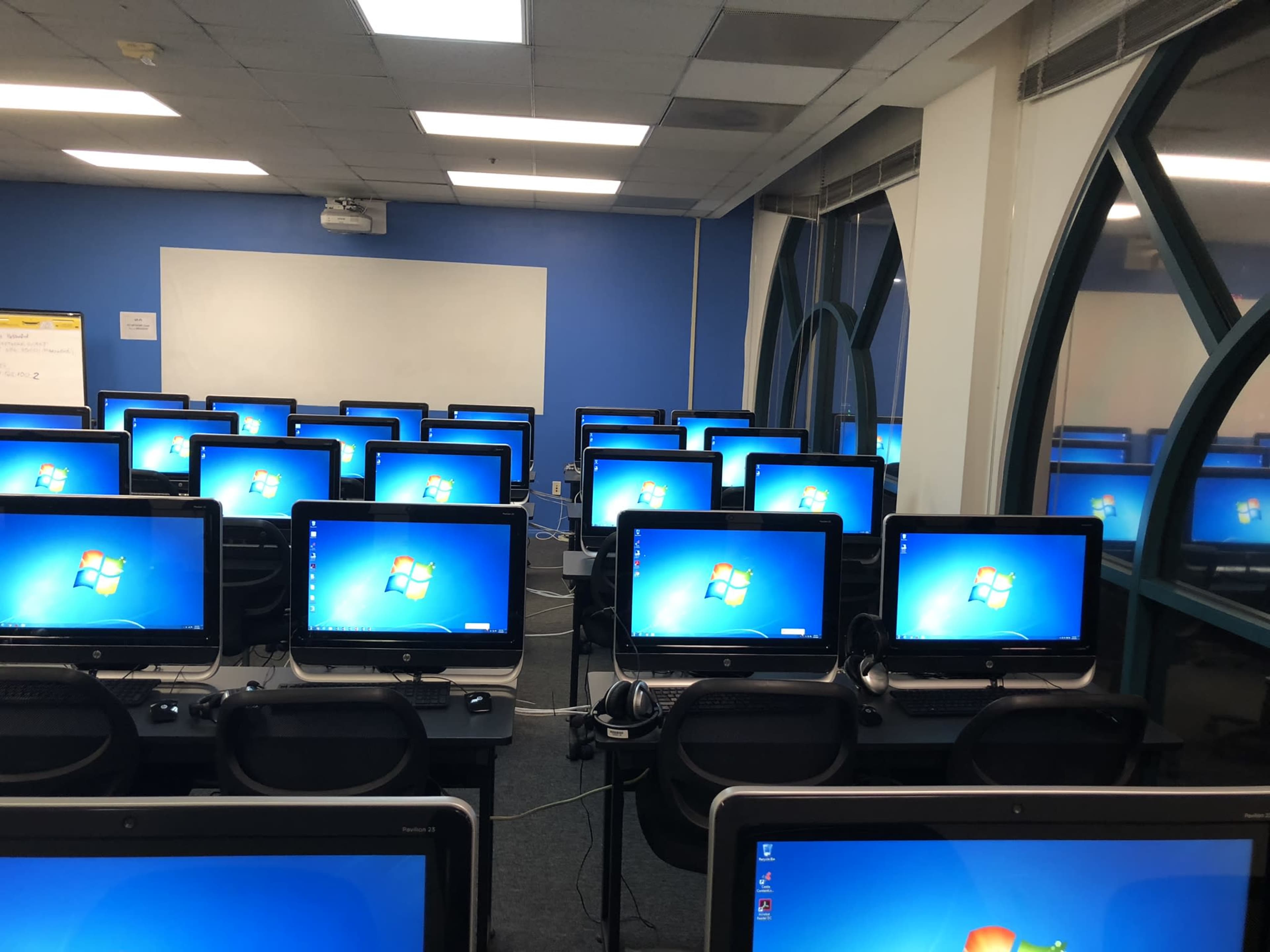 The image shows a room filled with multiple computer desks, each equipped with a monitor displaying the Windows desktop.