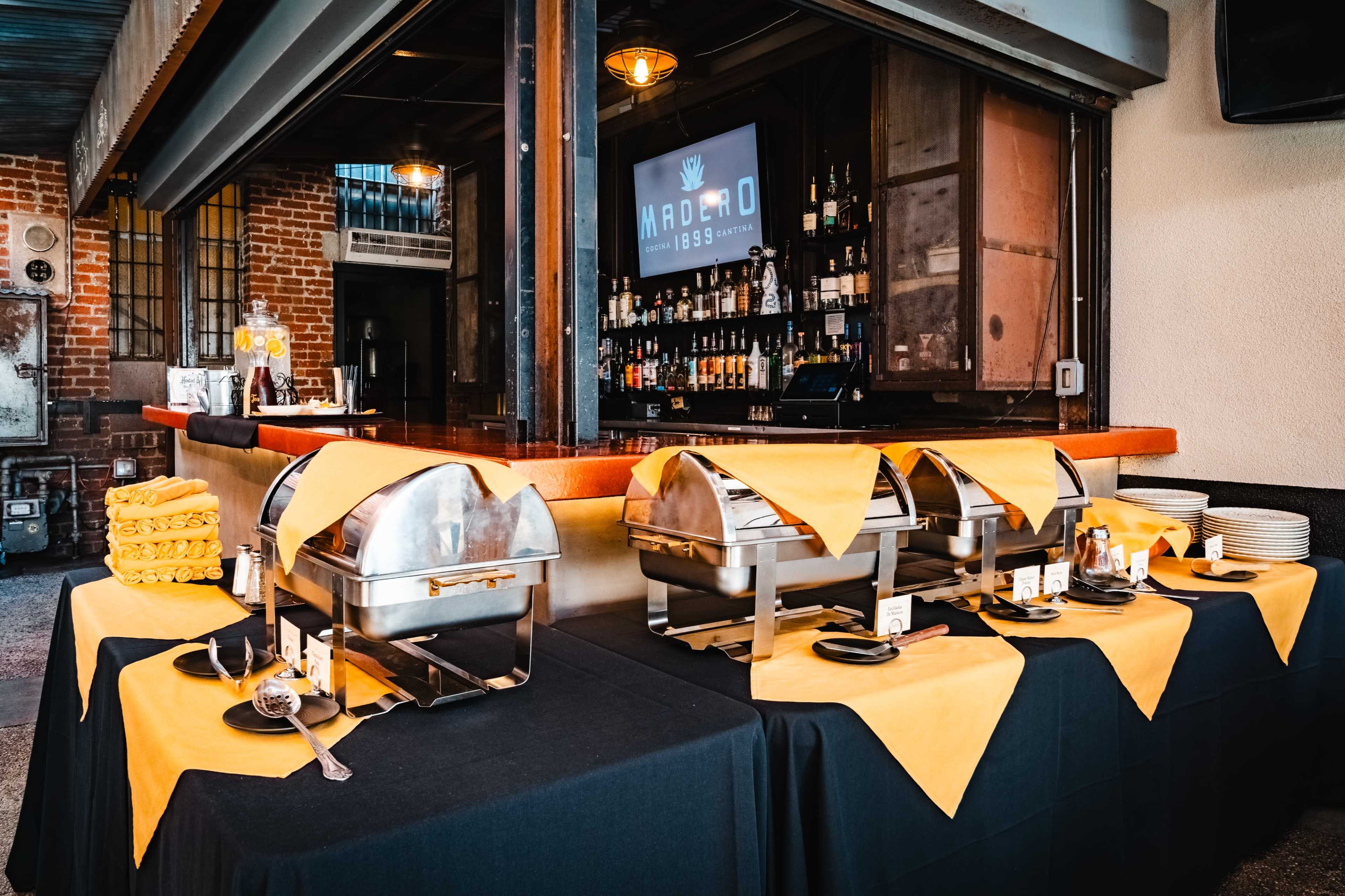 The image shows a buffet setup with chafing dishes on black tables adorned with yellow tablecloths, near a bar area with various bottles displayed in the background.