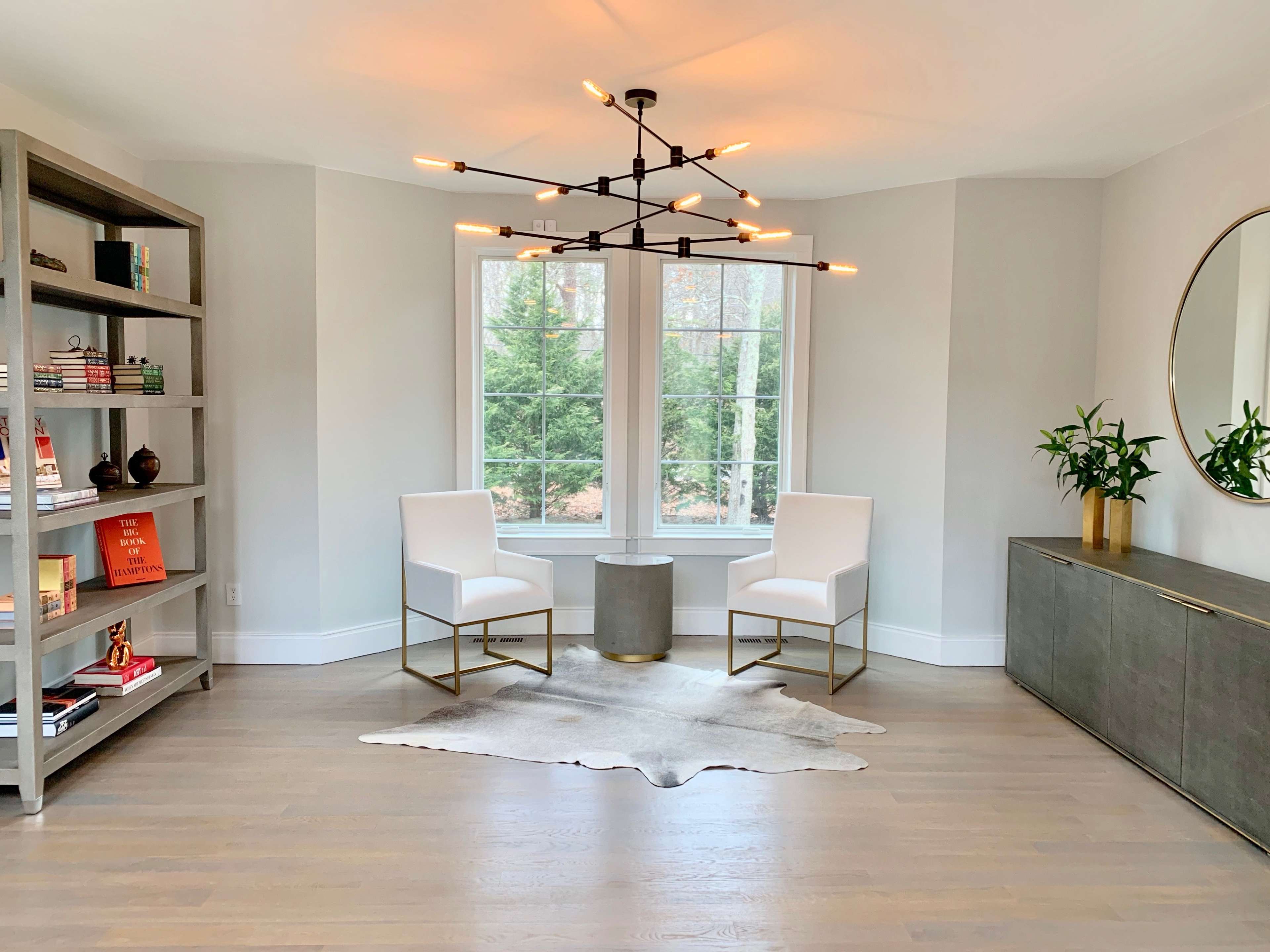 A modern sitting area features two white chairs, a round table, and a bookshelf against a wall with large windows providing natural light.