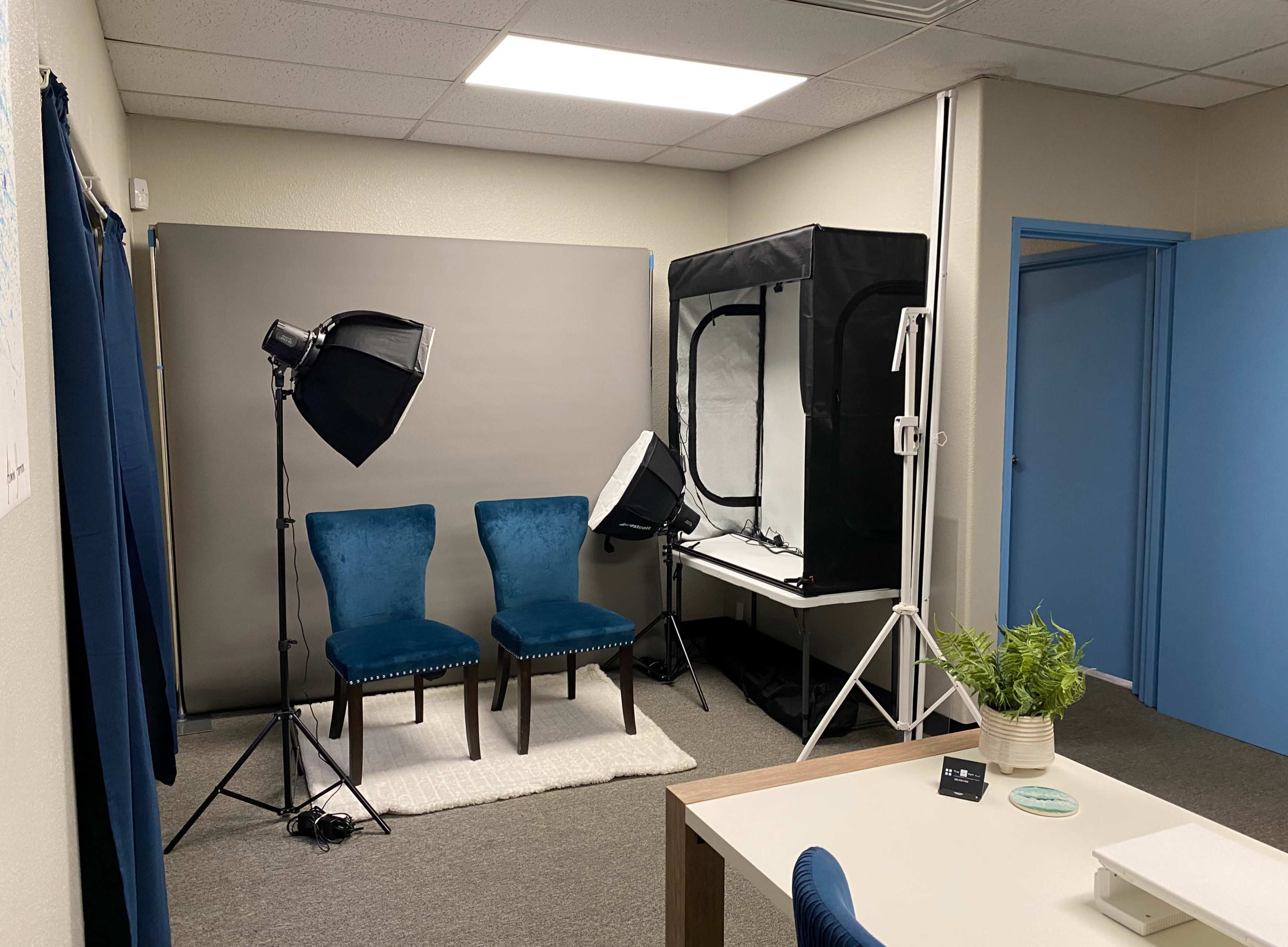 The image shows a small photography studio with two blue chairs, a light setup, and a backdrop in a well-lit room.