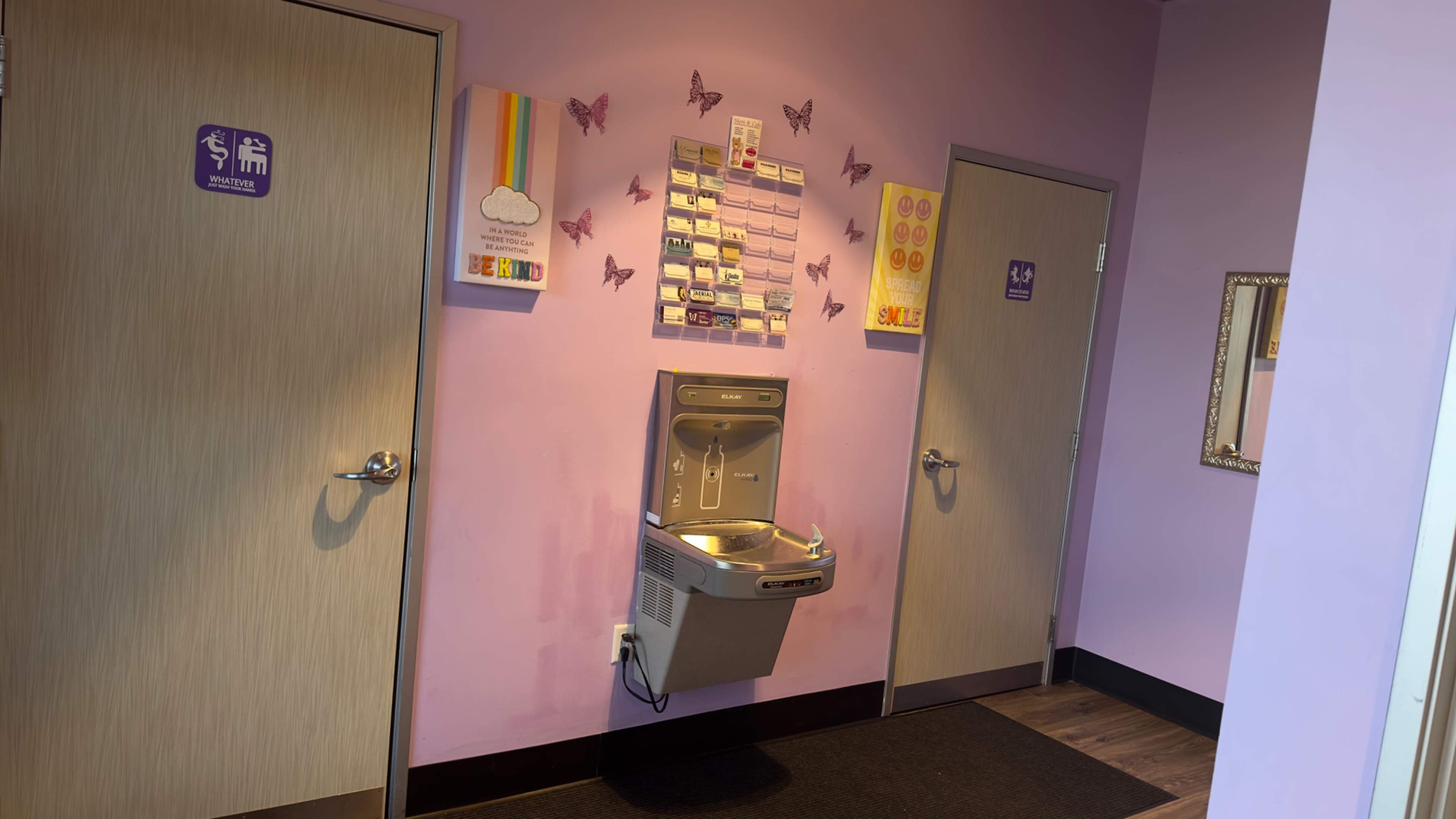 A hallway with a drinking fountain, two restroom doors, and colorful wall decorations featuring butterflies and various posters on a light purple wall.