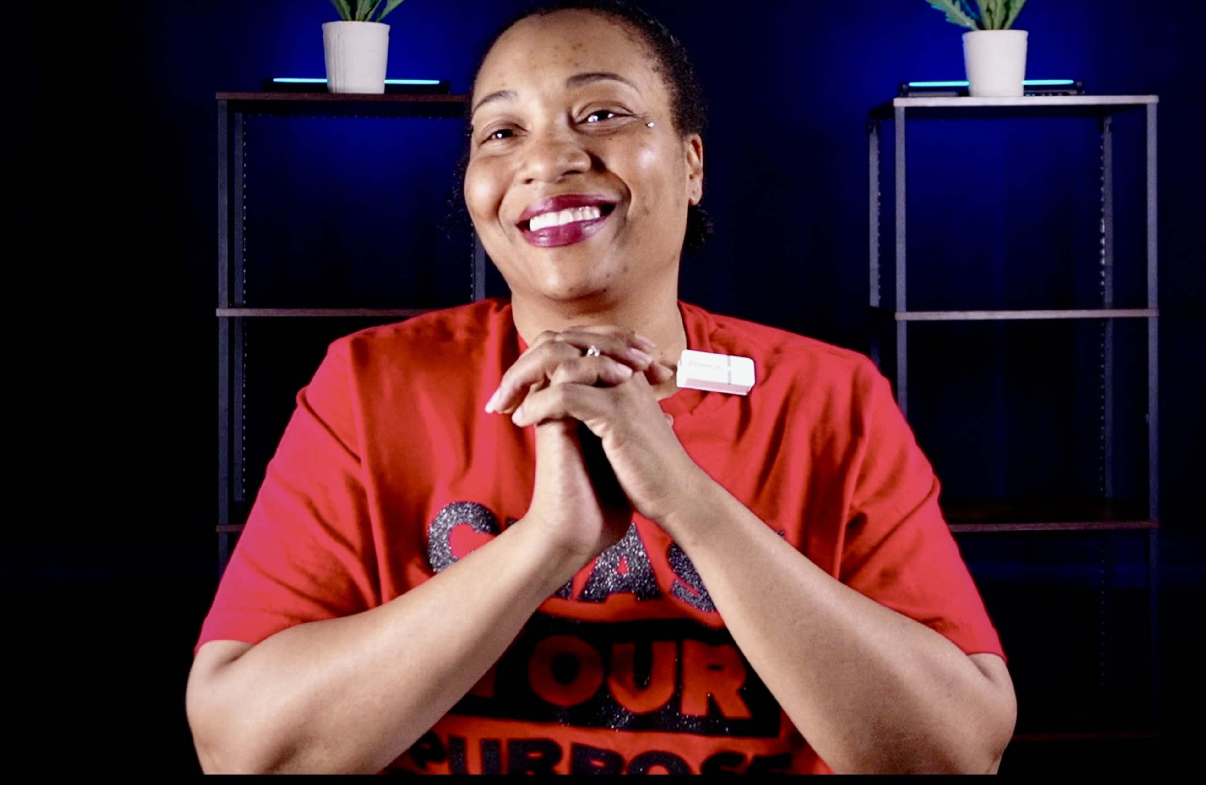 A woman in a red shirt is smiling and clasping her hands together in front of a dark background with shelves and potted plants.