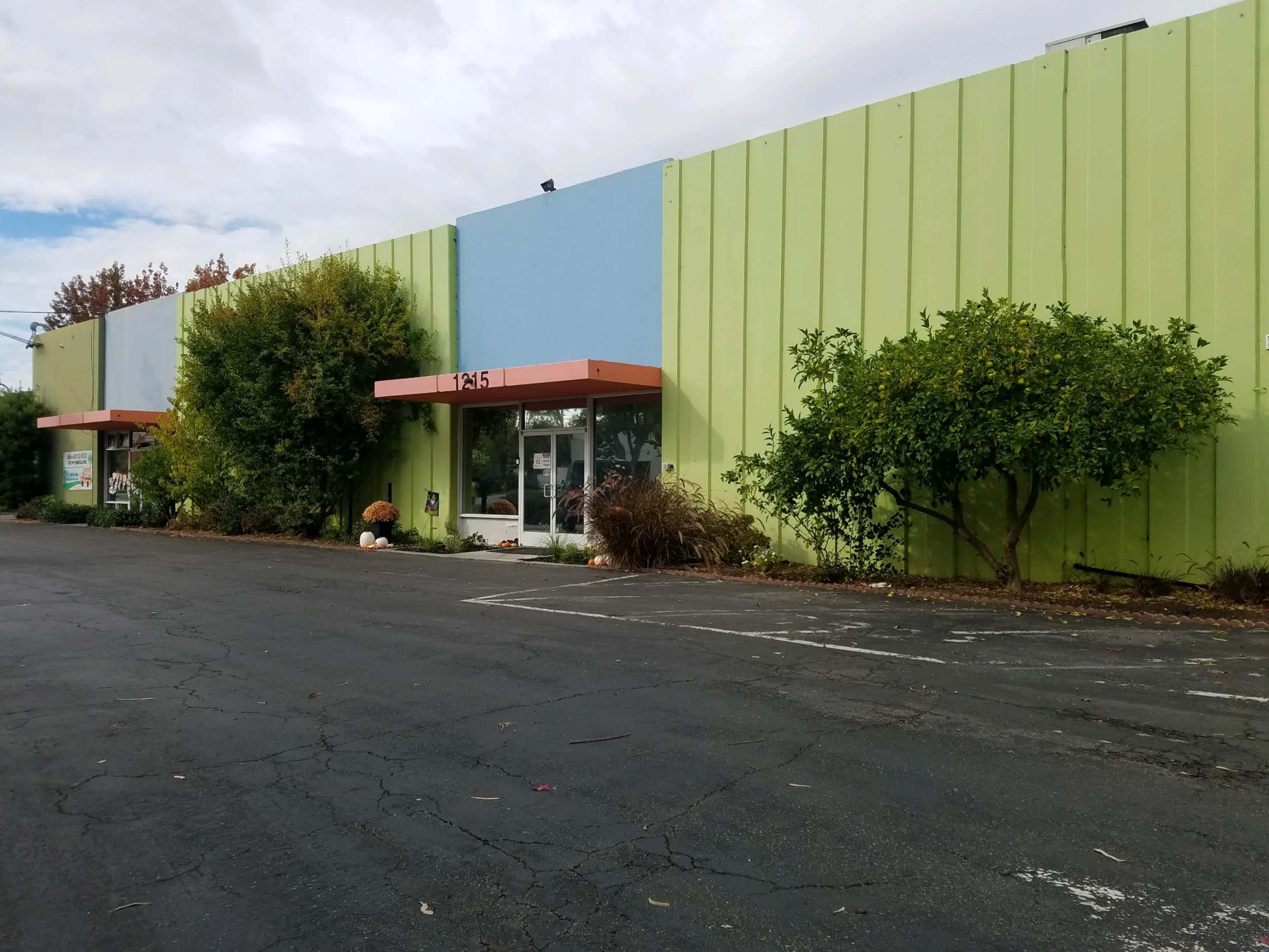 A single-story commercial building with a green and blue exterior, surrounded by shrubs and trees, and a cracked asphalt parking lot in front.