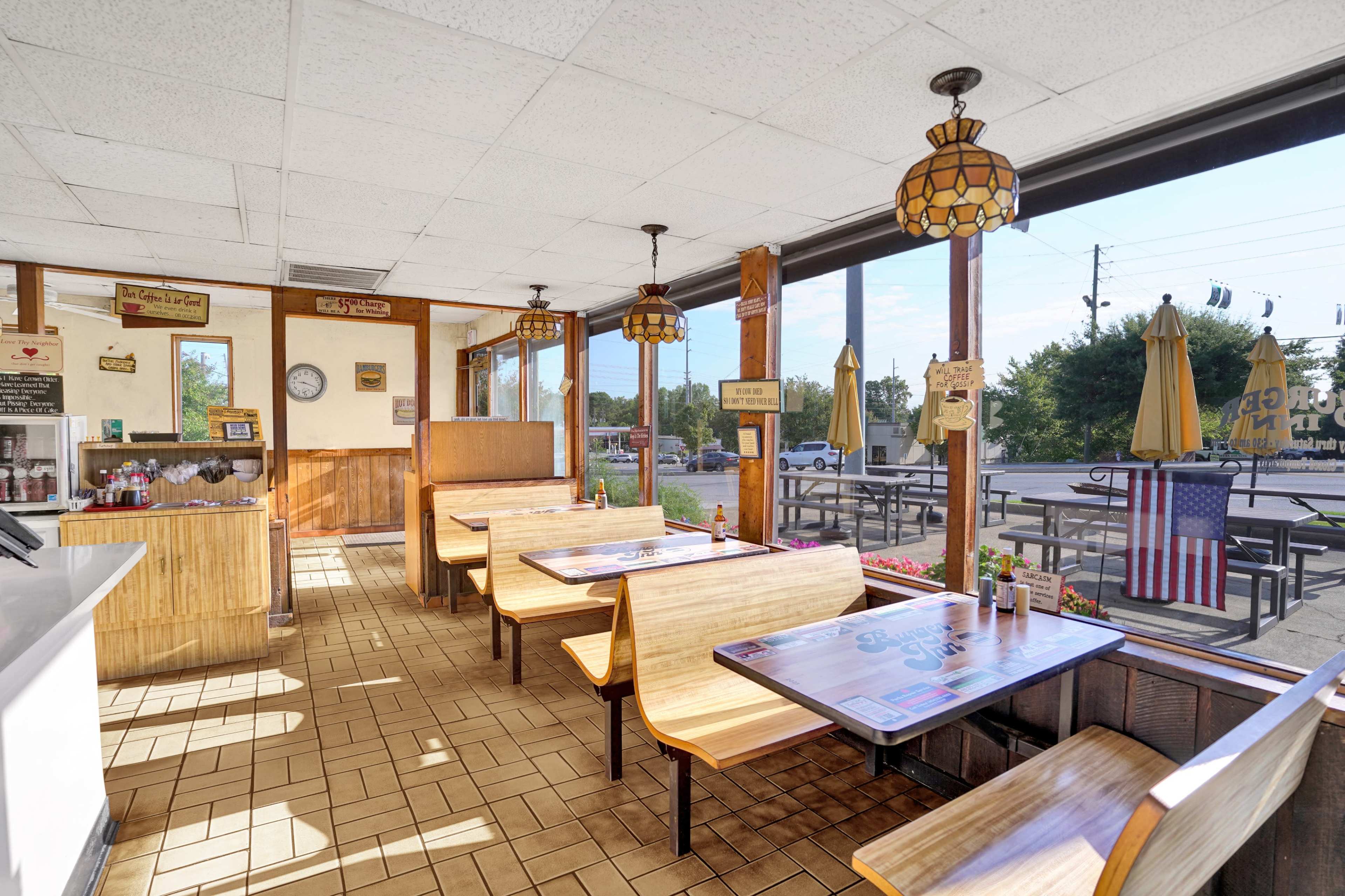 The image shows the interior of a diner with wooden booths, tables, and hanging pendant lights, featuring large windows that offer a view of the outdoor seating area.