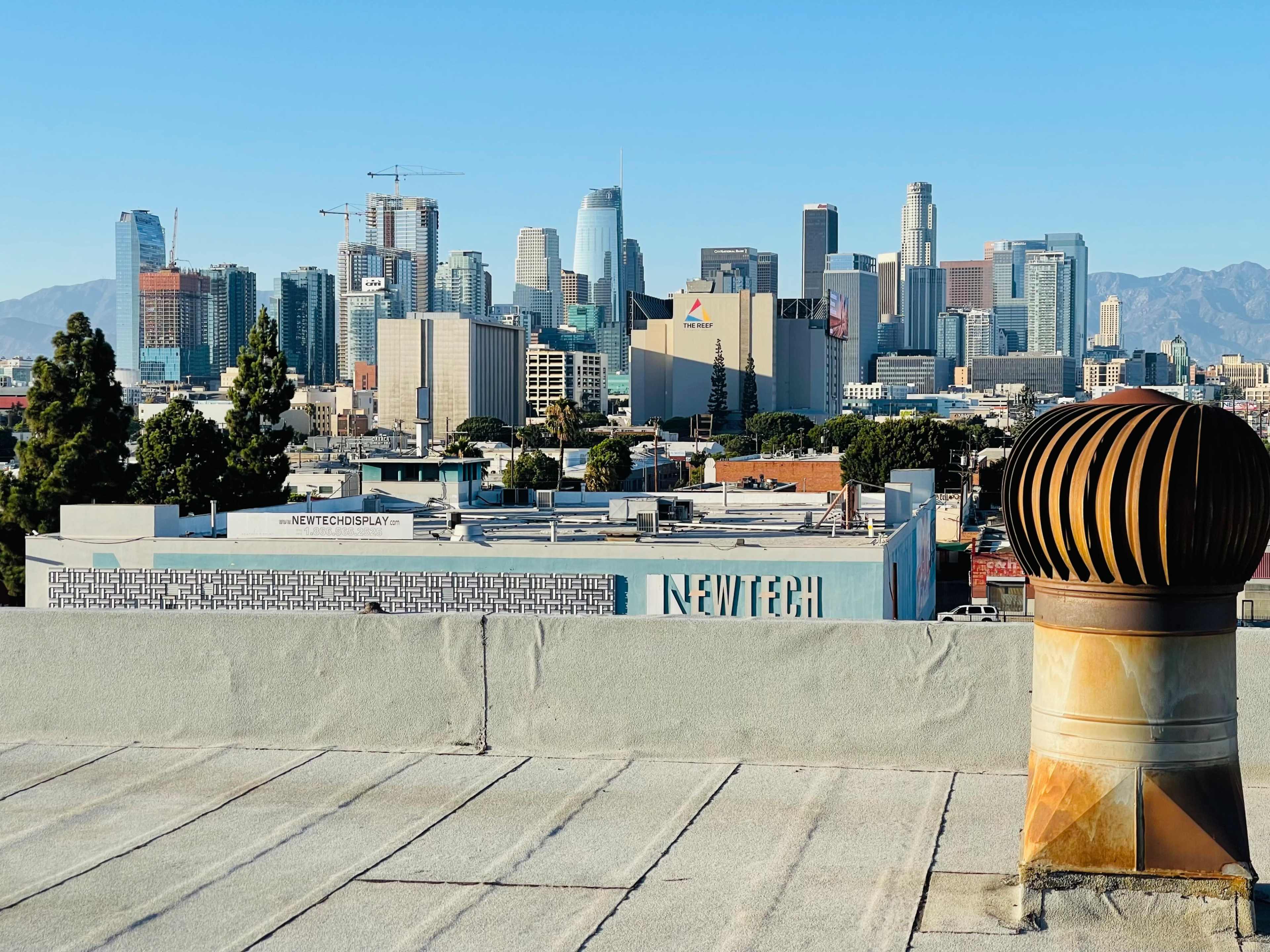 A rooftop view overlooking the Los Angeles skyline, featuring various high-rise buildings and a distant mountain range.