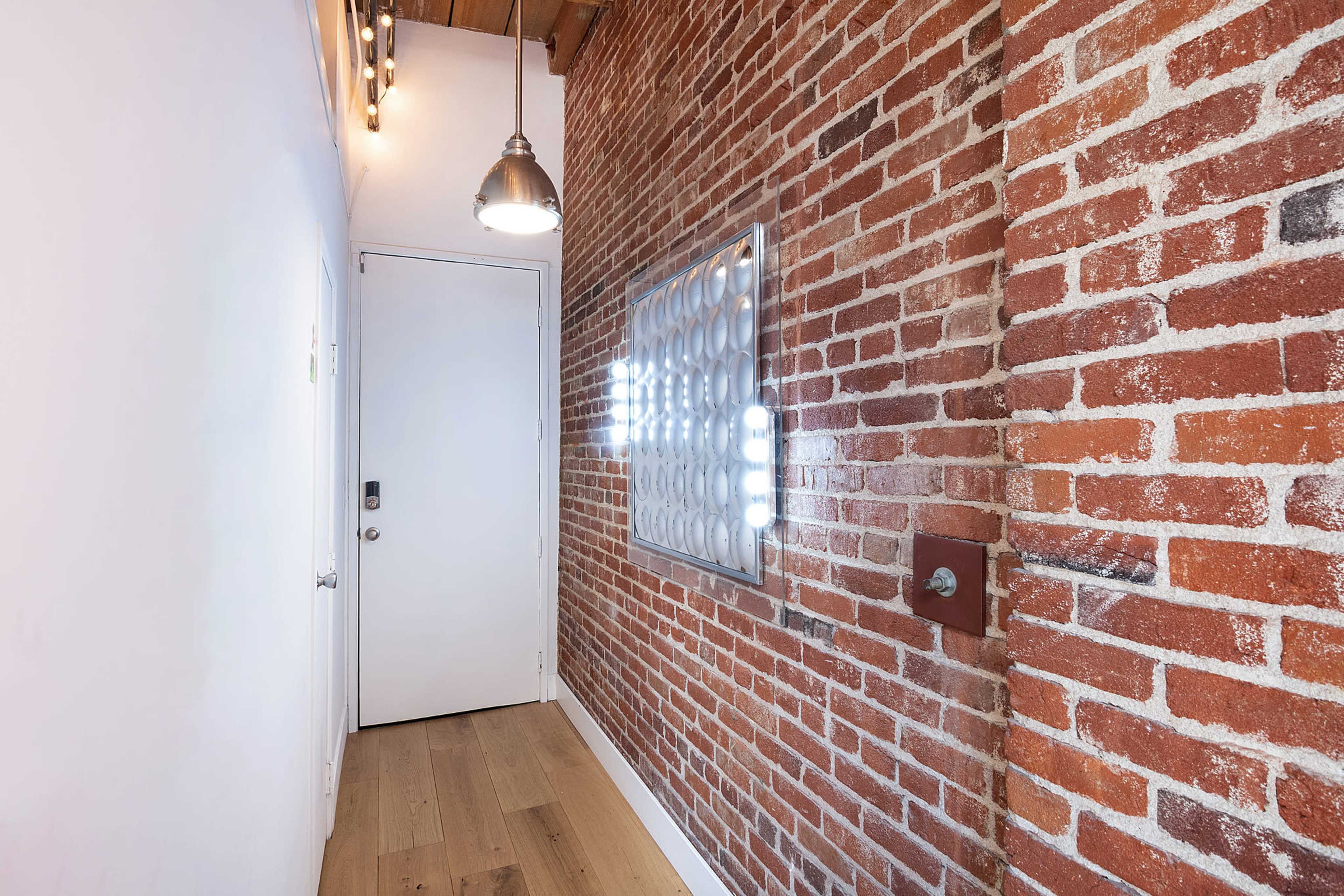 A hallway features a textured red brick wall opposite a smooth white wall, with a door at the end and a unique wall-mounted light fixture.