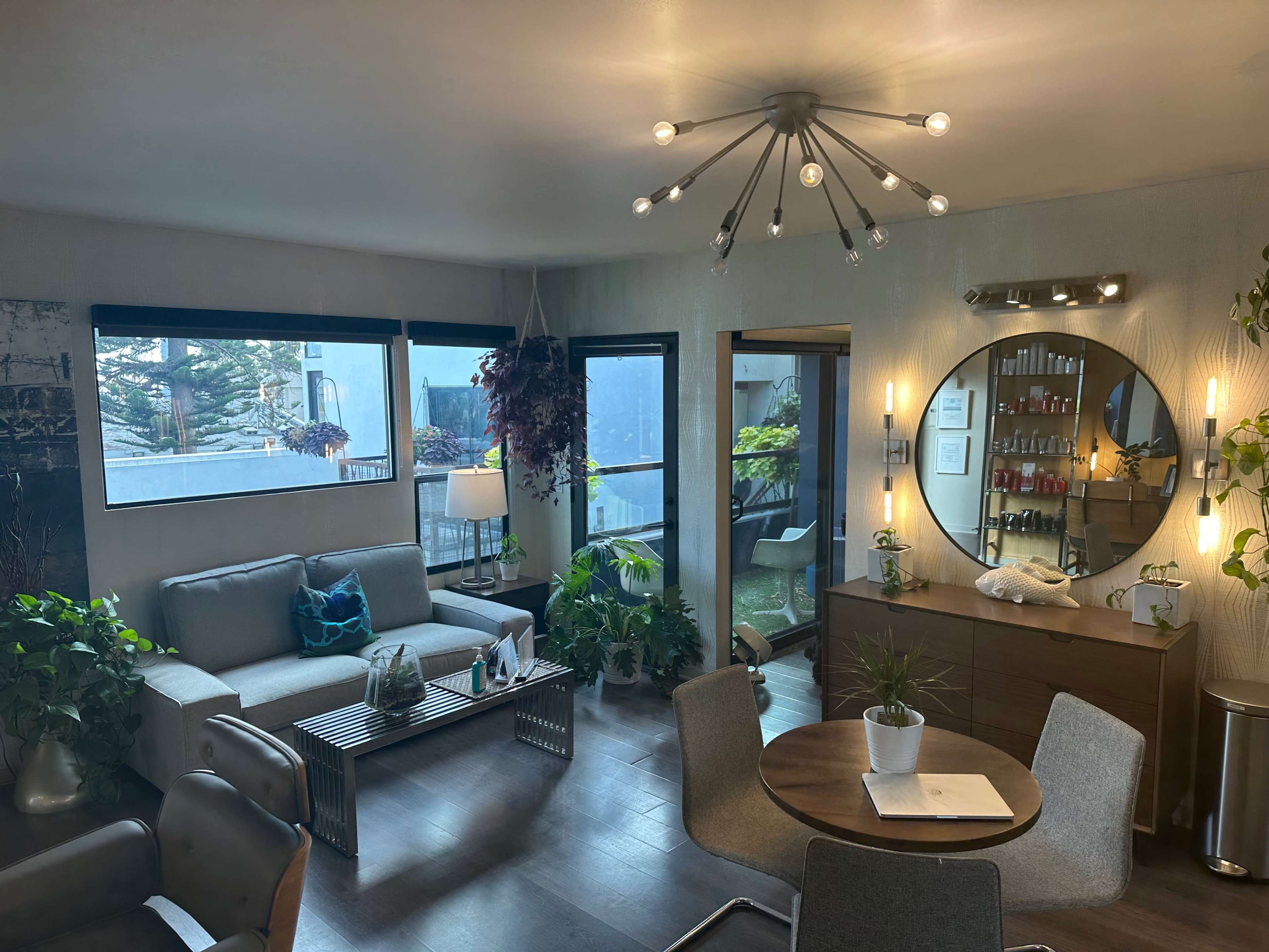 A modern living space features a gray sofa, a round dining table, and large windows allowing natural light, surrounded by indoor plants.