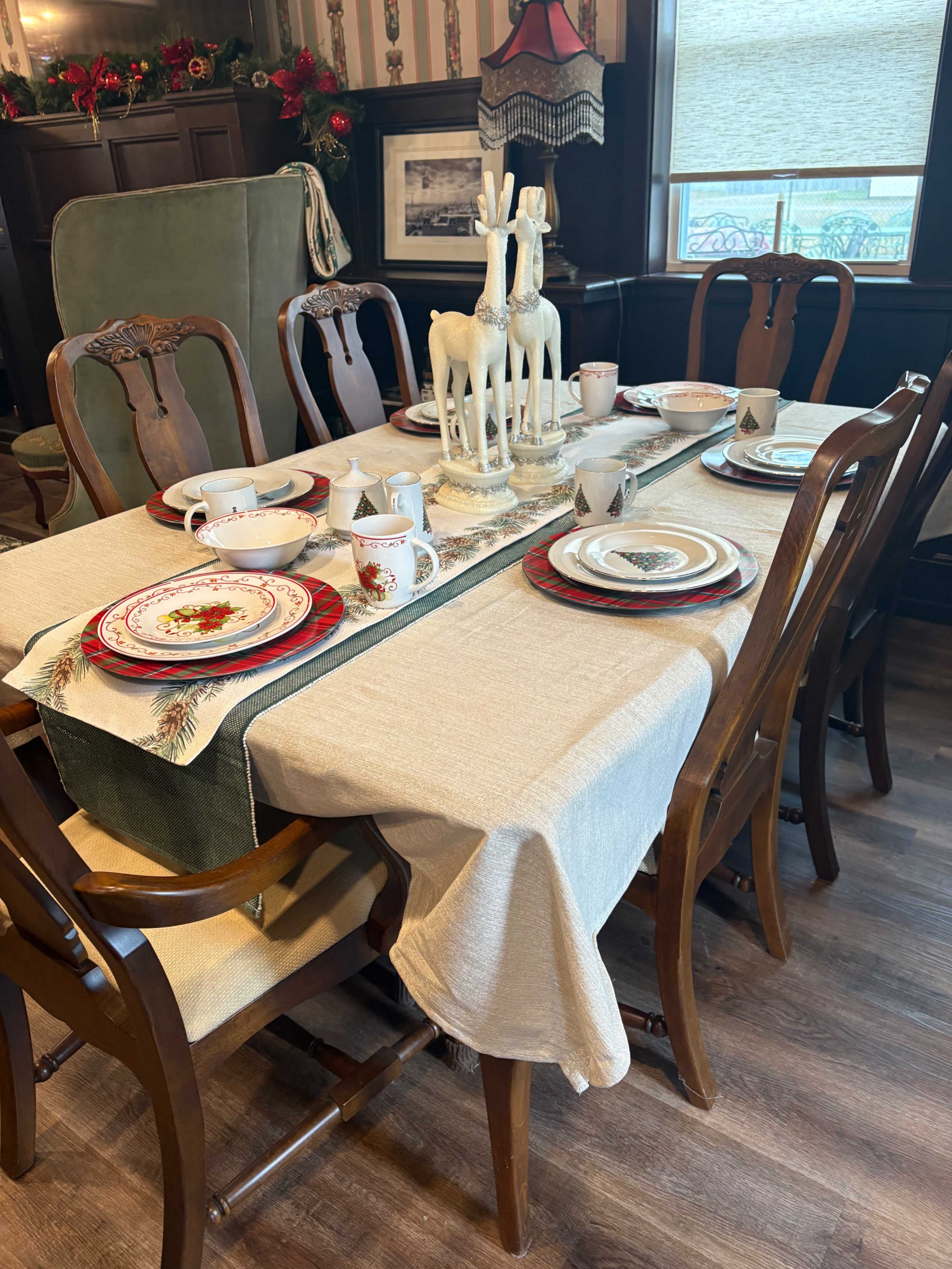 A dining table is set with a festive tablecloth, plates, and beverages, featuring decorative reindeer figurines and holiday-themed dishes in a cozy room.