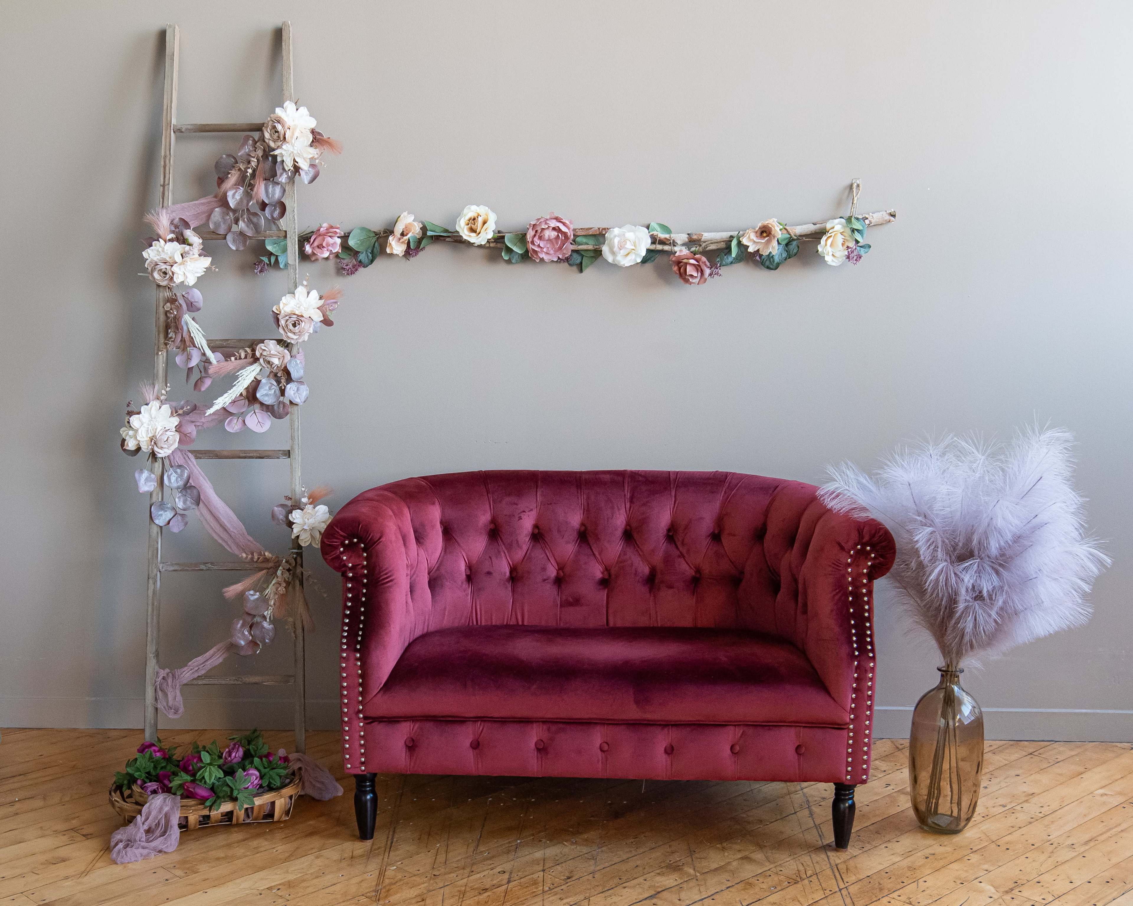 The image features a plush, burgundy tufted sofa positioned in front of a light gray wall, accompanied by a decorative ladder adorned with flowers and a tall vase holding soft purple pampas grass.